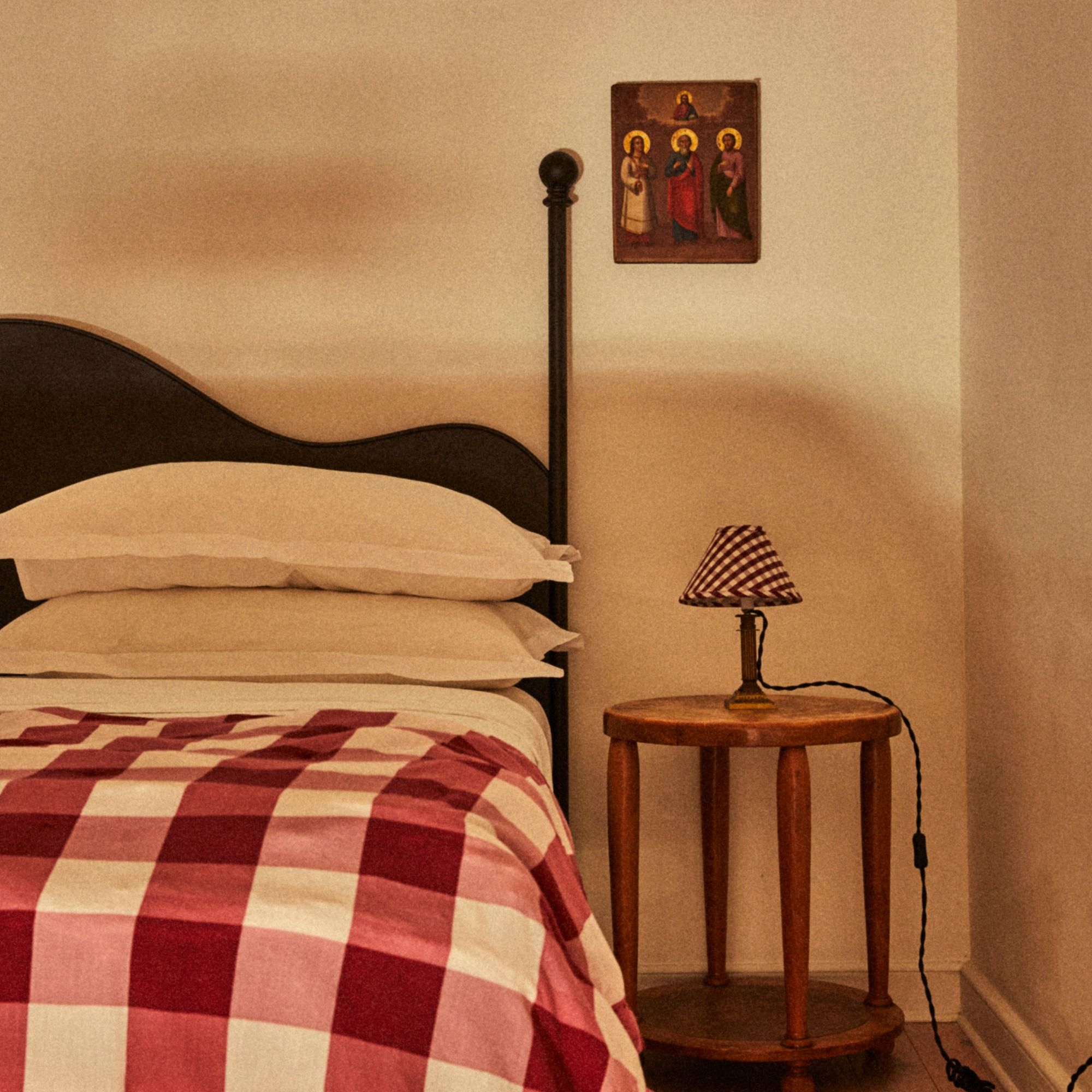 Cozy bedroom scene with a bed covered in a red and white checkered blanket, a black headboard, a small wooden side table, a gingham lamp, and a wall painting.