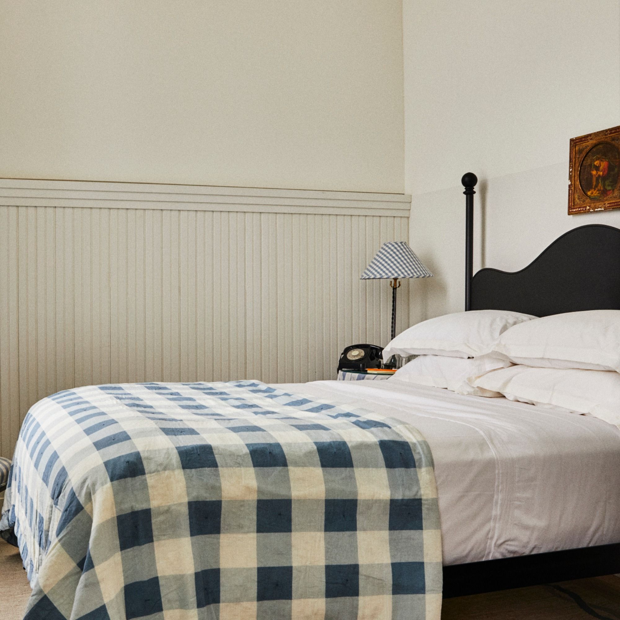 A cozy bedroom with a dark headboard, white bedding, and a blue-checkered blanket. A vintage phone and lamp sit on the side table.
