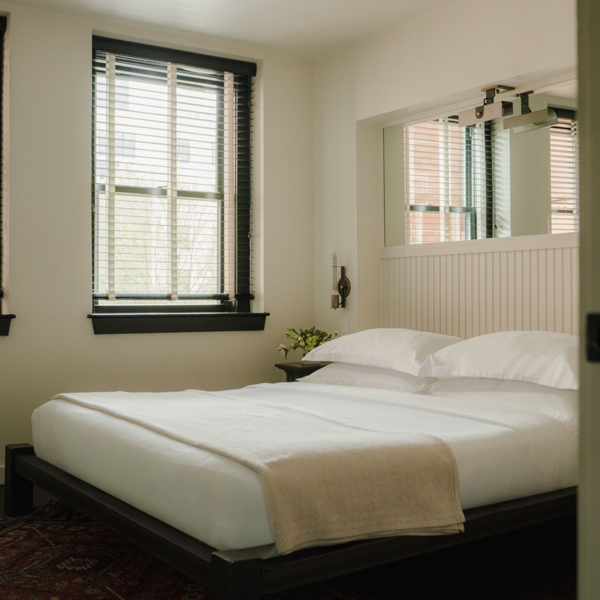A cozy bedroom with a neatly made double bed, white linens, a large window with blinds, and a small bedside table with a plant.