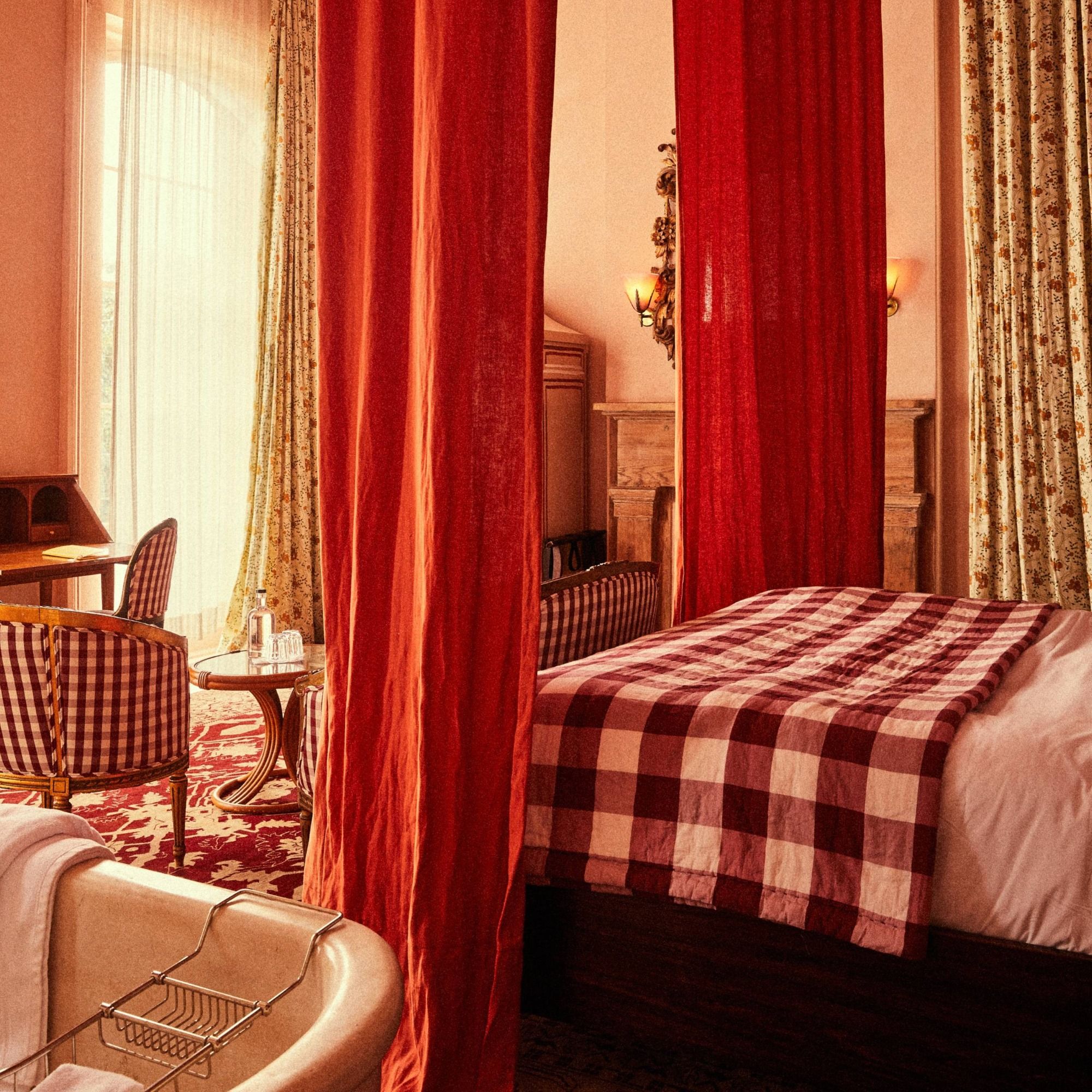 Cozy bedroom with red curtains, checkered bedding, a clawfoot bathtub, plaid chairs, and a sunlit window with floral drapes.