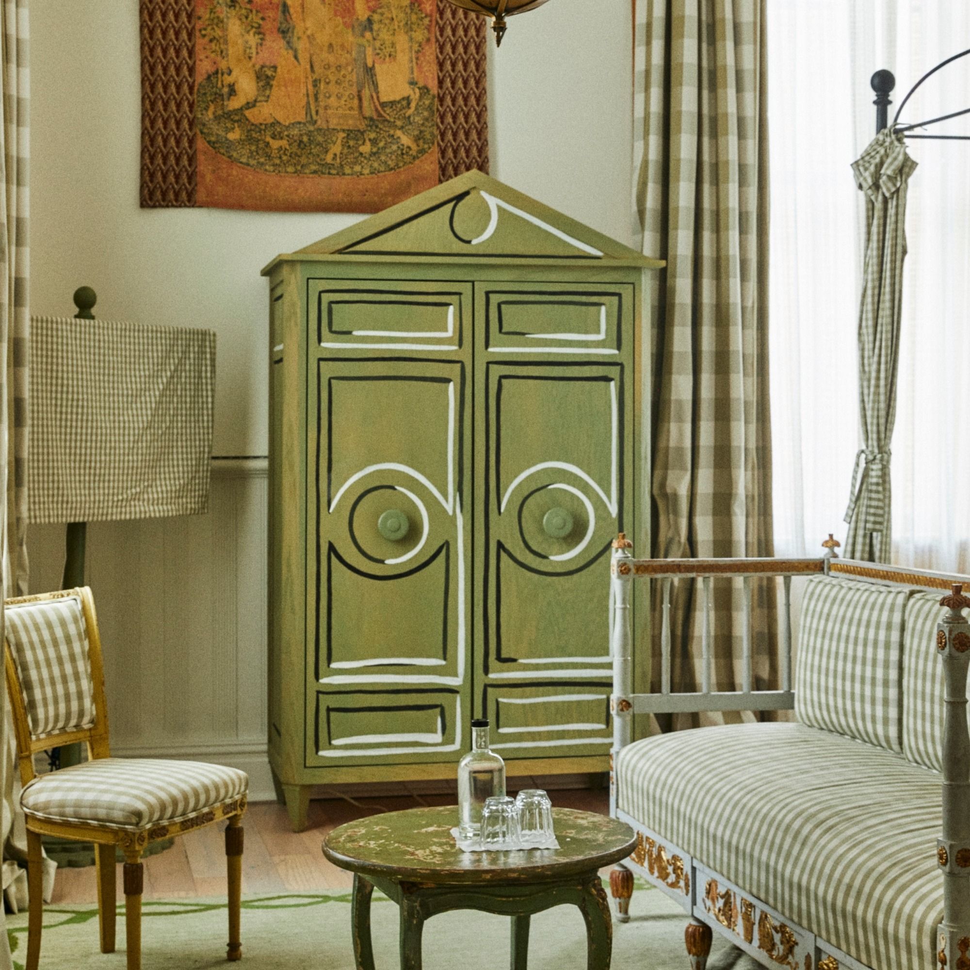 A cozy room with green checkered furniture, a decorative wardrobe, and a small round table with a bottle and glasses. Wall tapestry in the background.