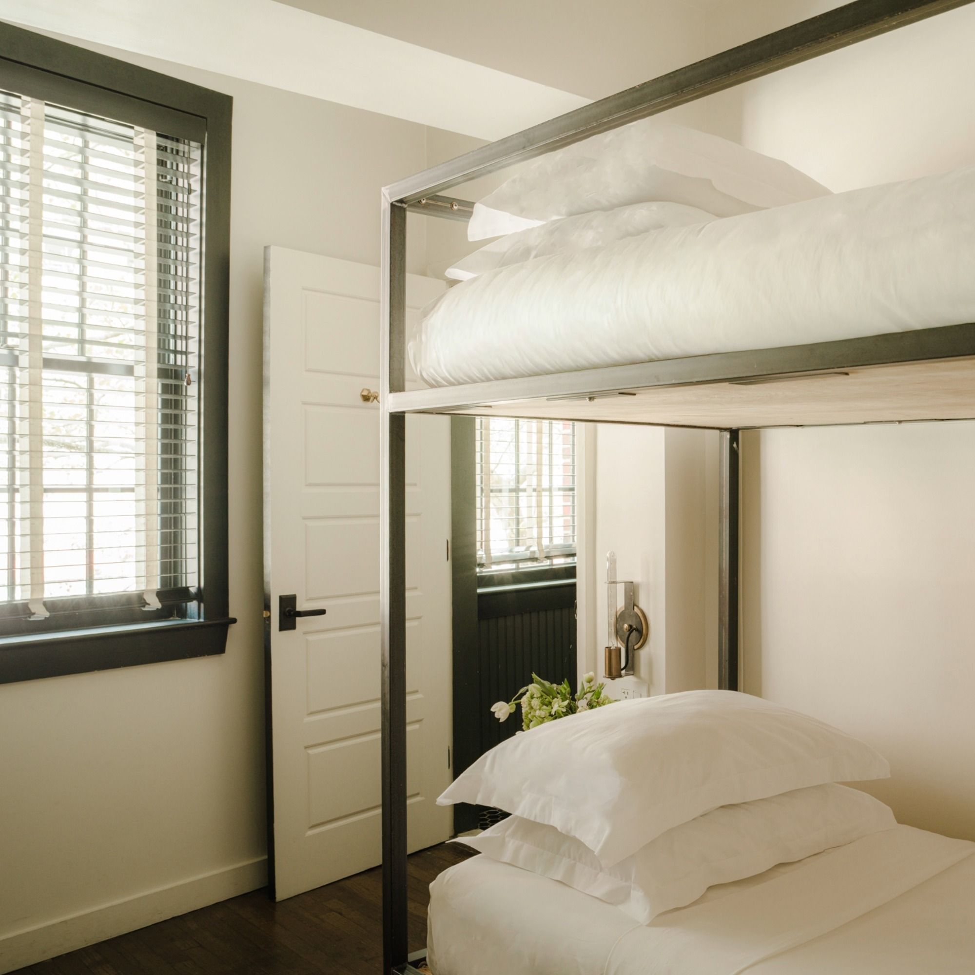 Minimalist bedroom with a modern bunk bed, white bedding, a small window with blinds, and a partially open door, creating an airy feel.