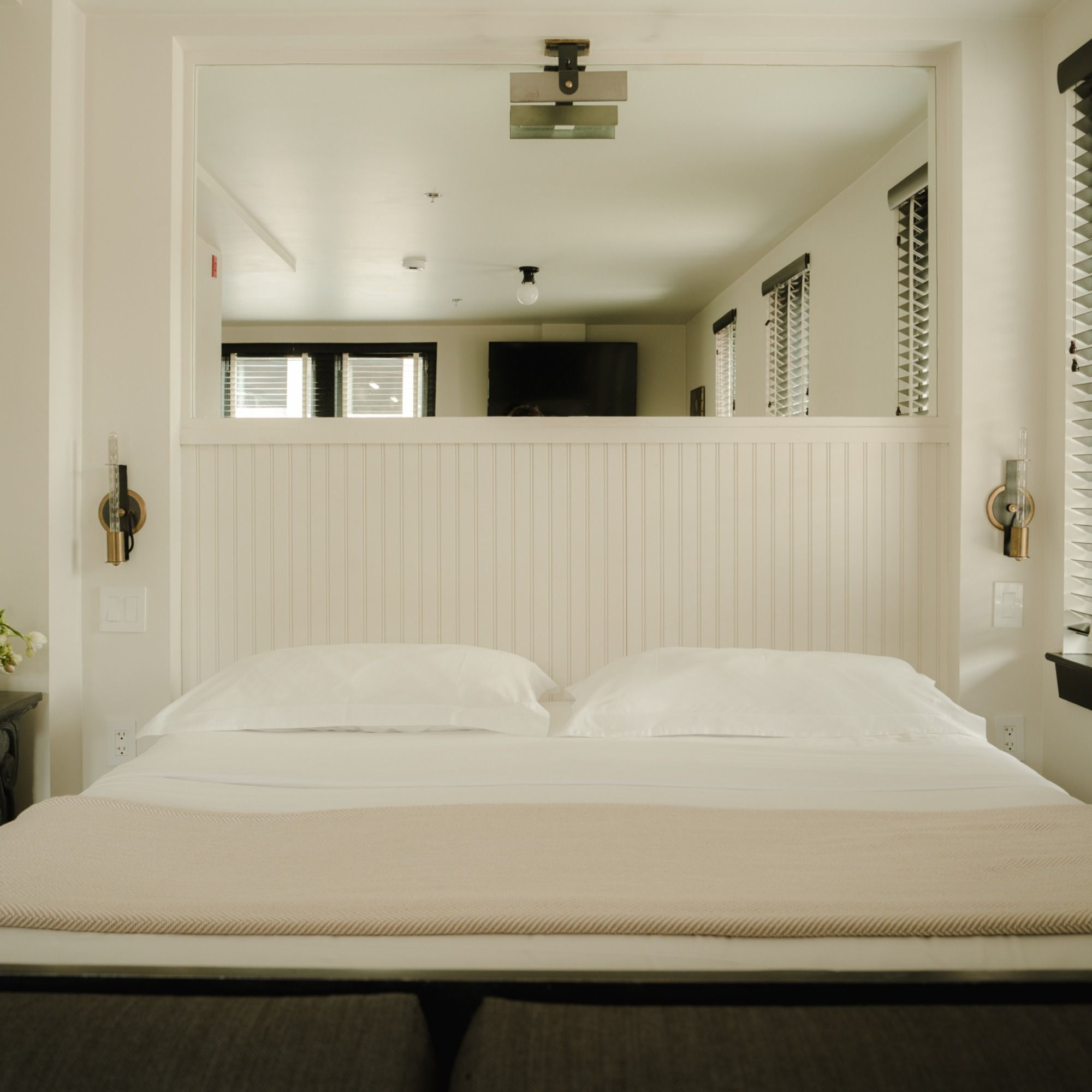 A neatly made bed with white pillows and a beige blanket in a well-lit, modern bedroom with white walls and windows with blinds.