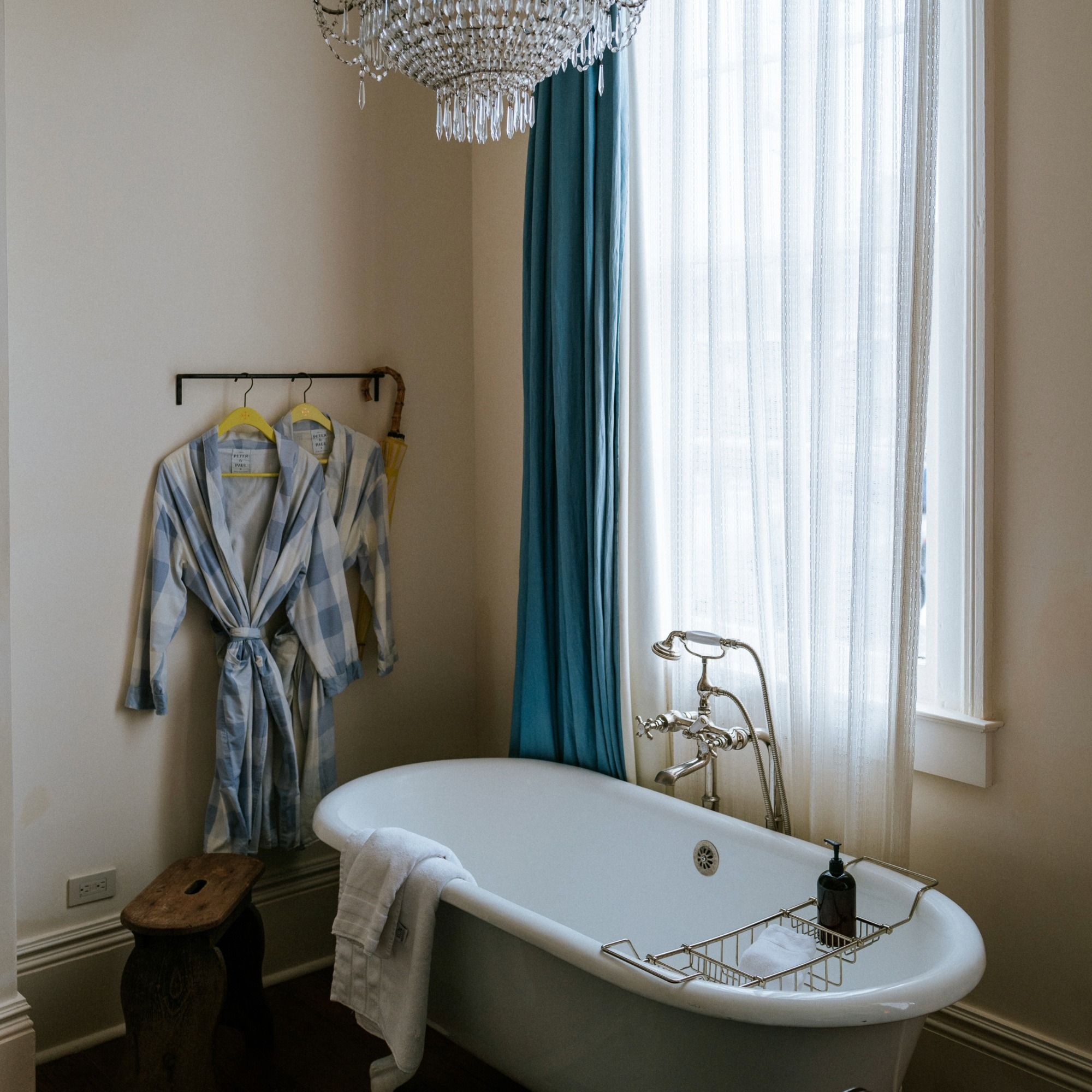 A cozy bathroom with a freestanding bathtub, hanging checkered bathrobe, wooden stool, chandelier, and blue curtains by a window.