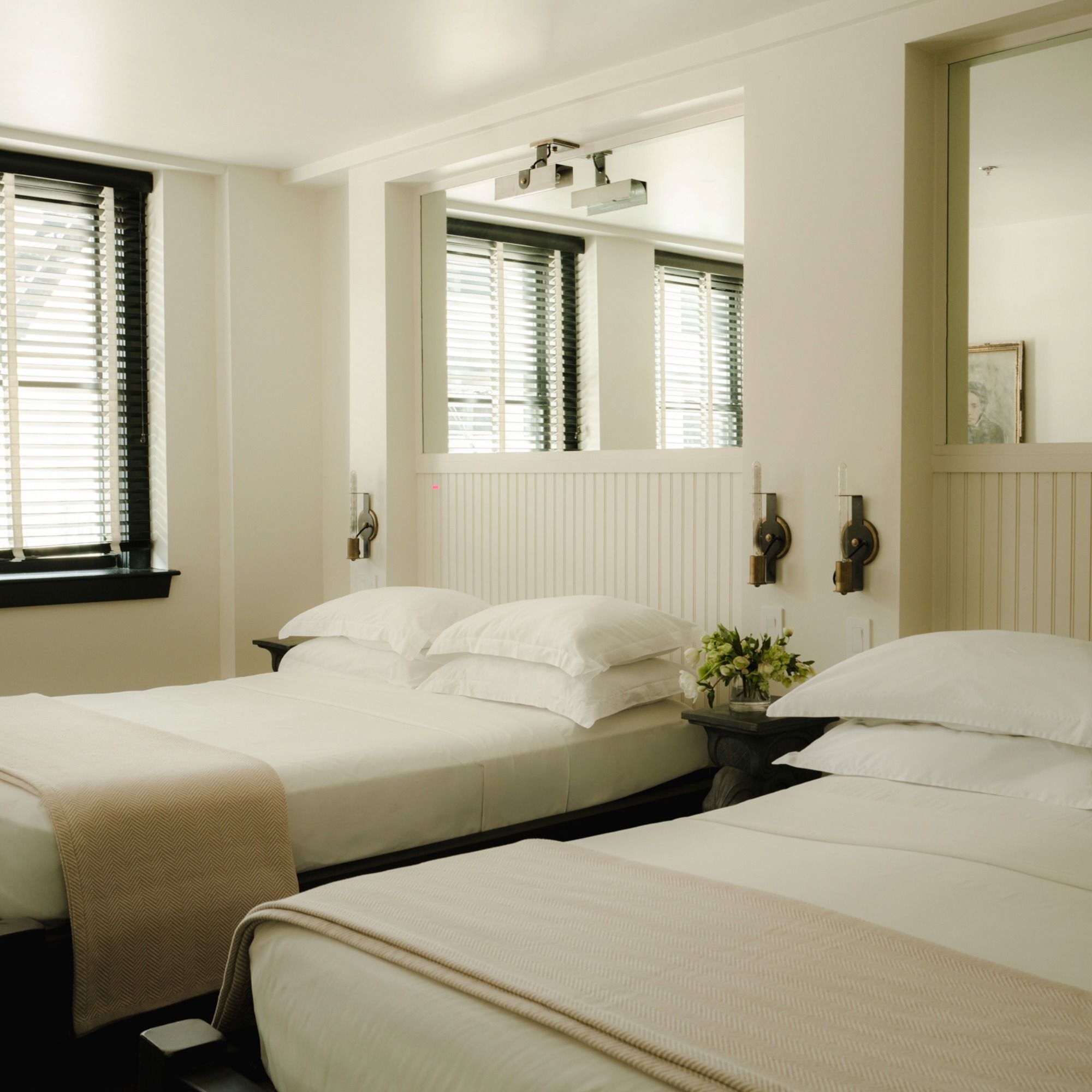 Two neatly made twin beds with white linens and beige throws in a bright room with a large mirror and shuttered windows.