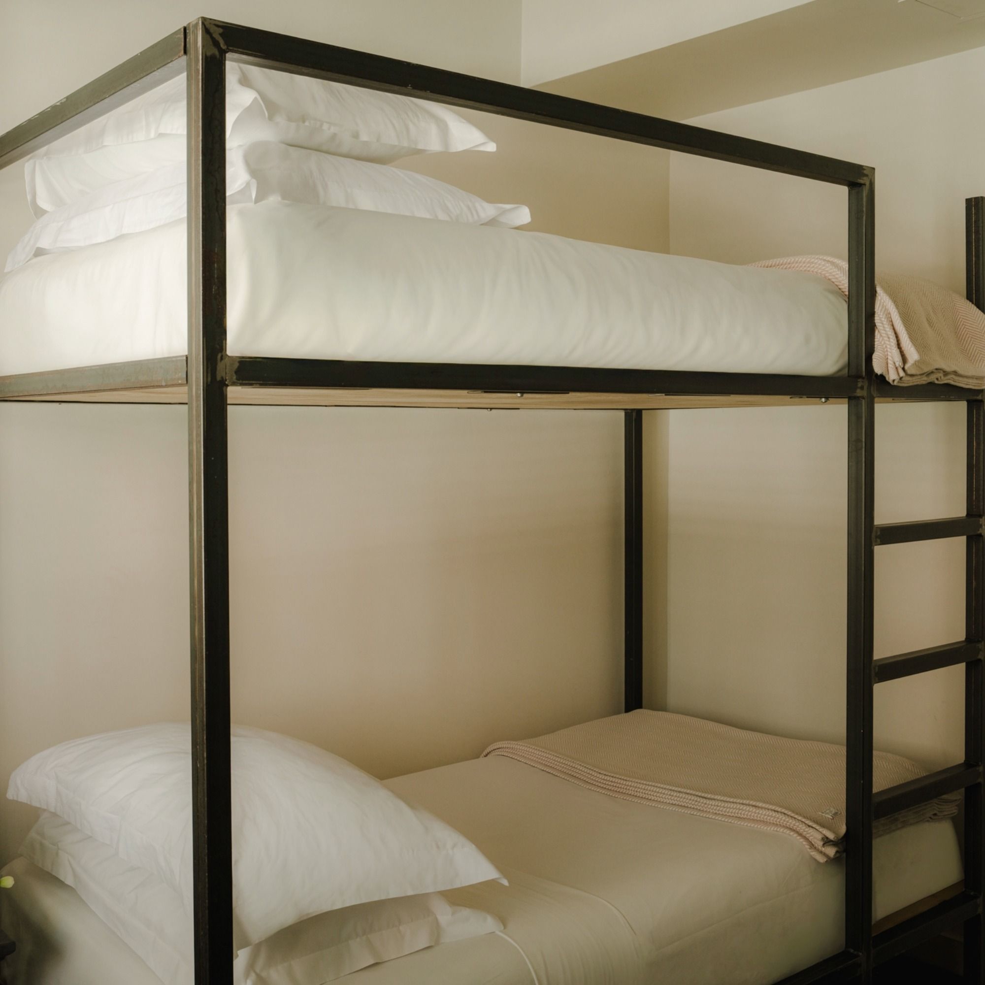 A minimalist metal-framed bunk bed with white bedding and pillows, and a folded beige blanket on the lower bunk.