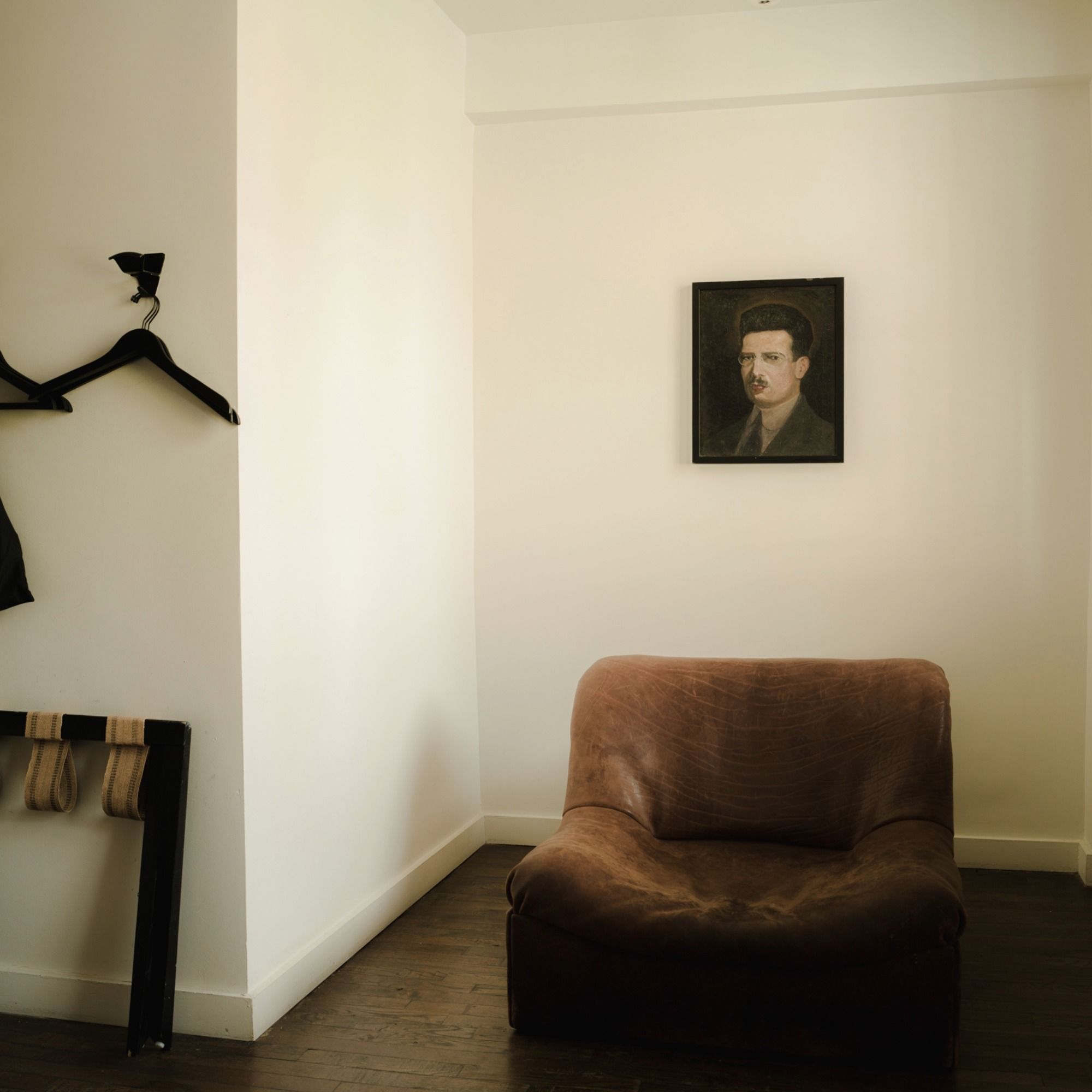A cozy room with a brown armchair, a wall-mounted portrait of a man, and coat hangers on a white wall with dark wooden flooring.