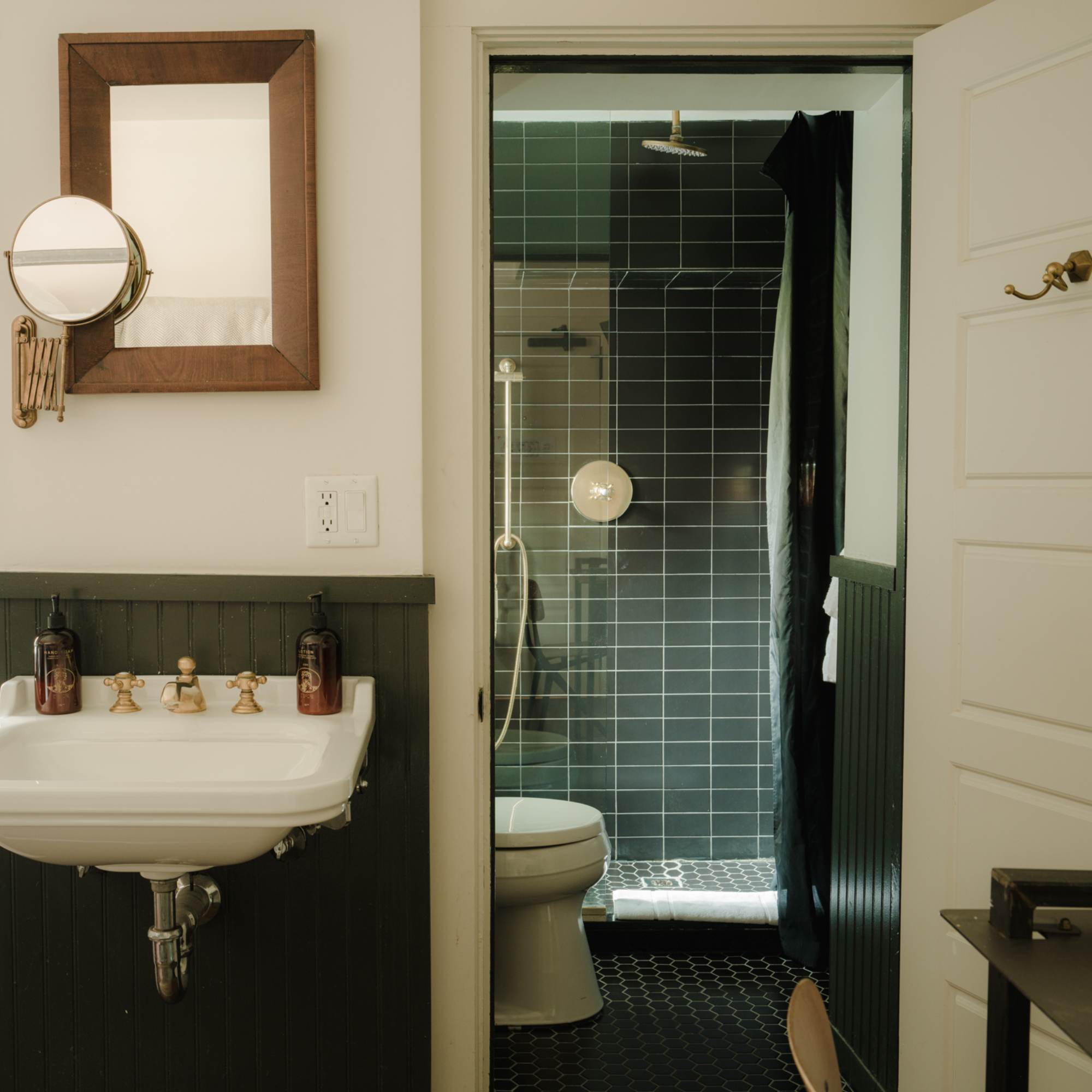A bathroom with a white sink, wooden mirror, black tile shower, black curtain, and toilet. Accessories on the sink include soap dispensers.