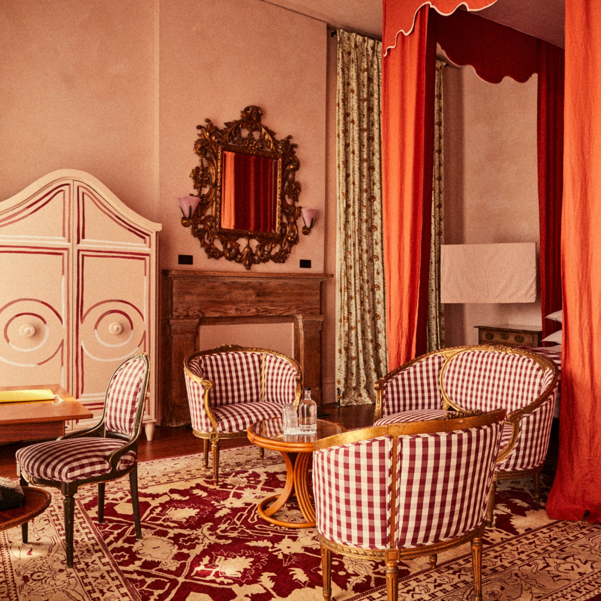 Vintage-style room with red checkered armchairs, ornate mirror, wooden fireplace, draped orange curtains, and a patterned carpet.