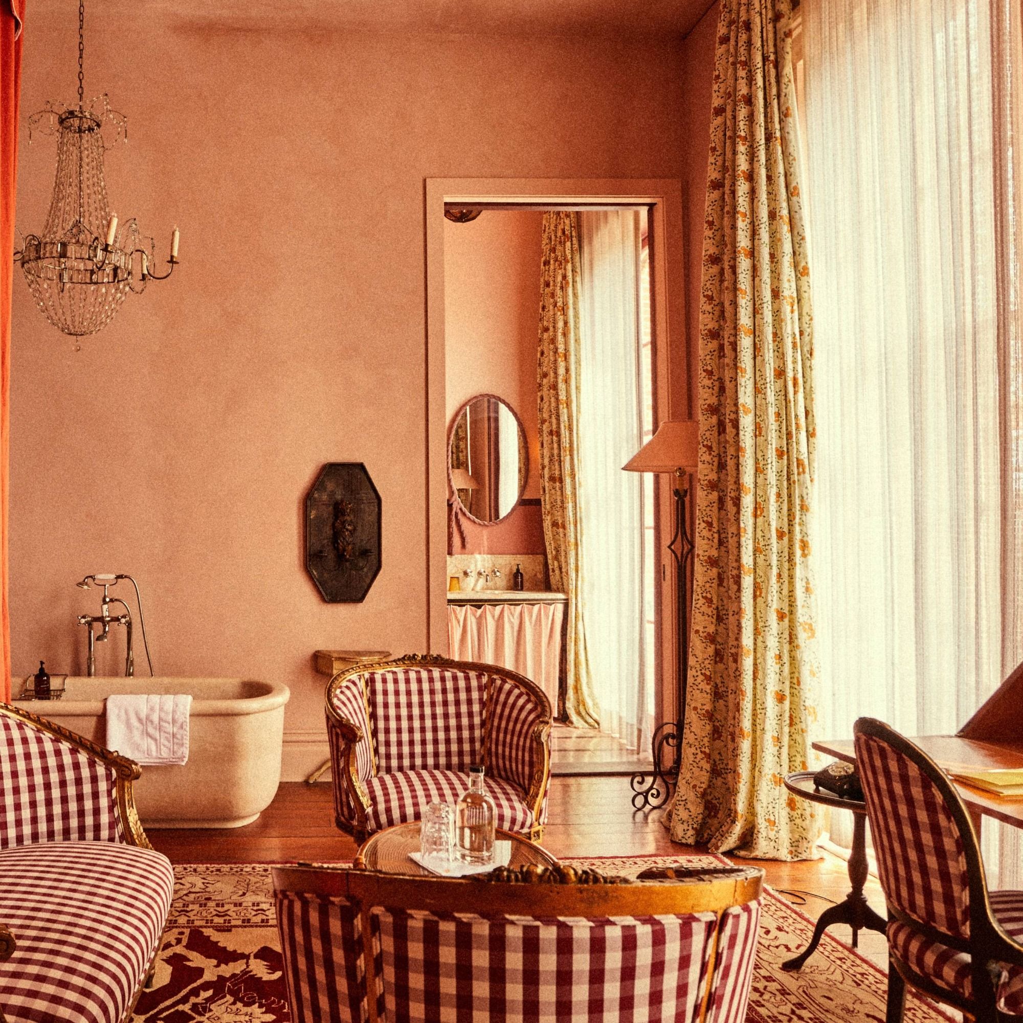Vintage-style room with pink walls, patterned curtains, checkered chairs, a clawfoot bathtub, chandelier, and ornate decor.