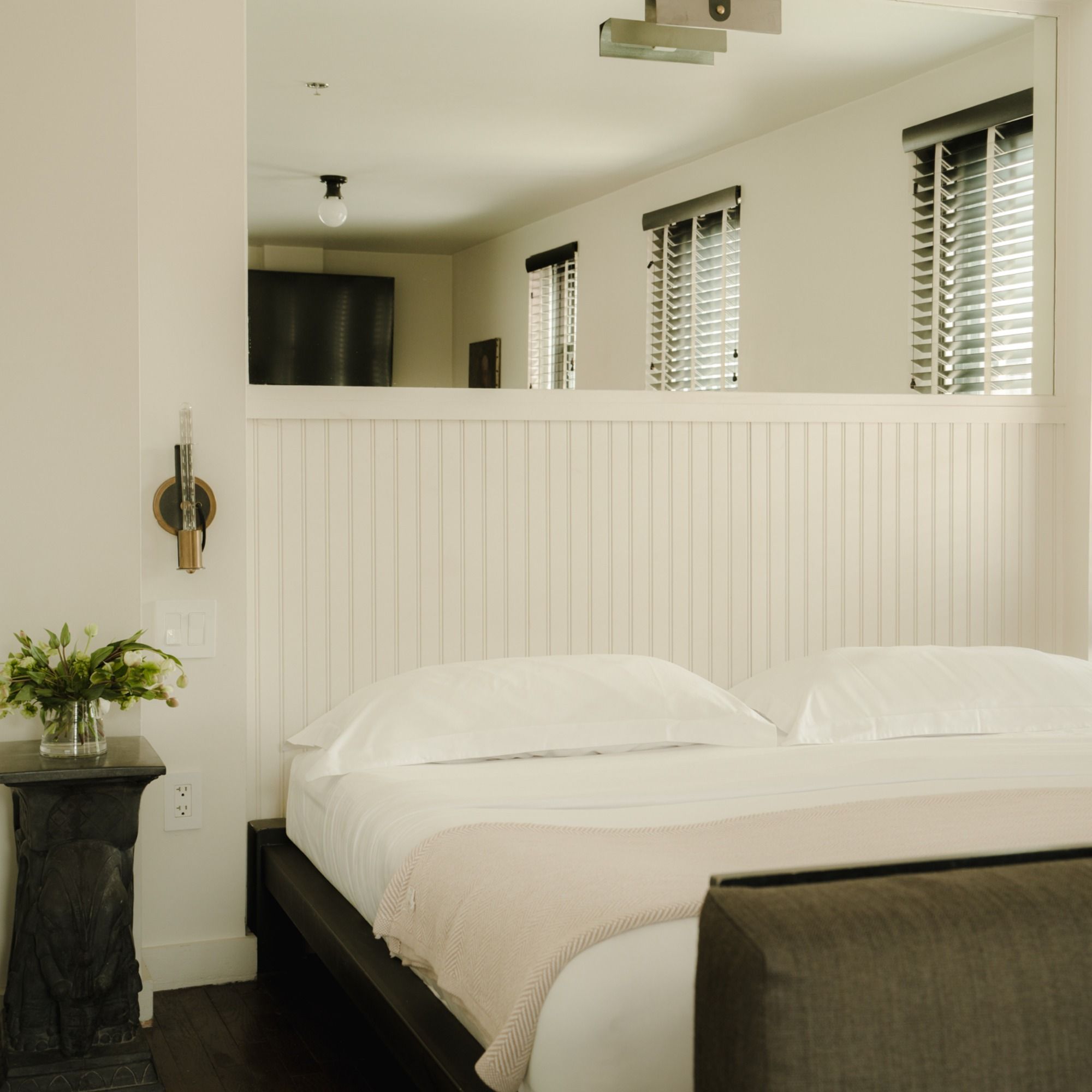 Minimalist bedroom with a white bed, a beige throw, a small side table with a vase of flowers, and a half-wall under windows with blinds.