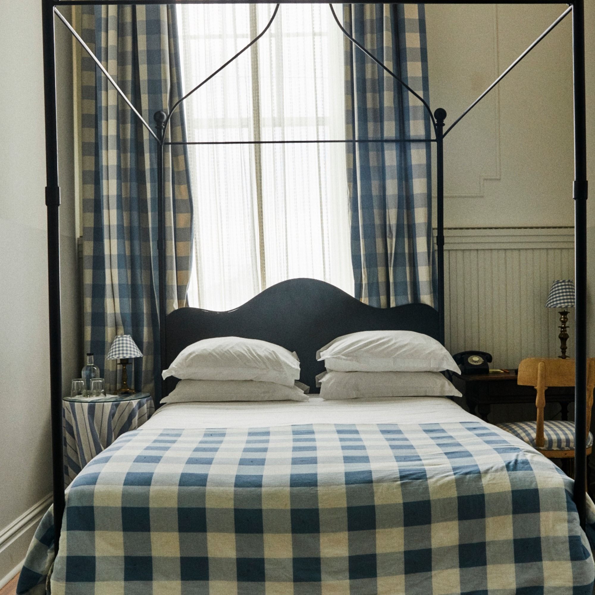 A cozy bedroom with a double bed covered in blue and white plaid bedding, matching curtains, and a black canopy frame. Vintage lamp and phone on side table.