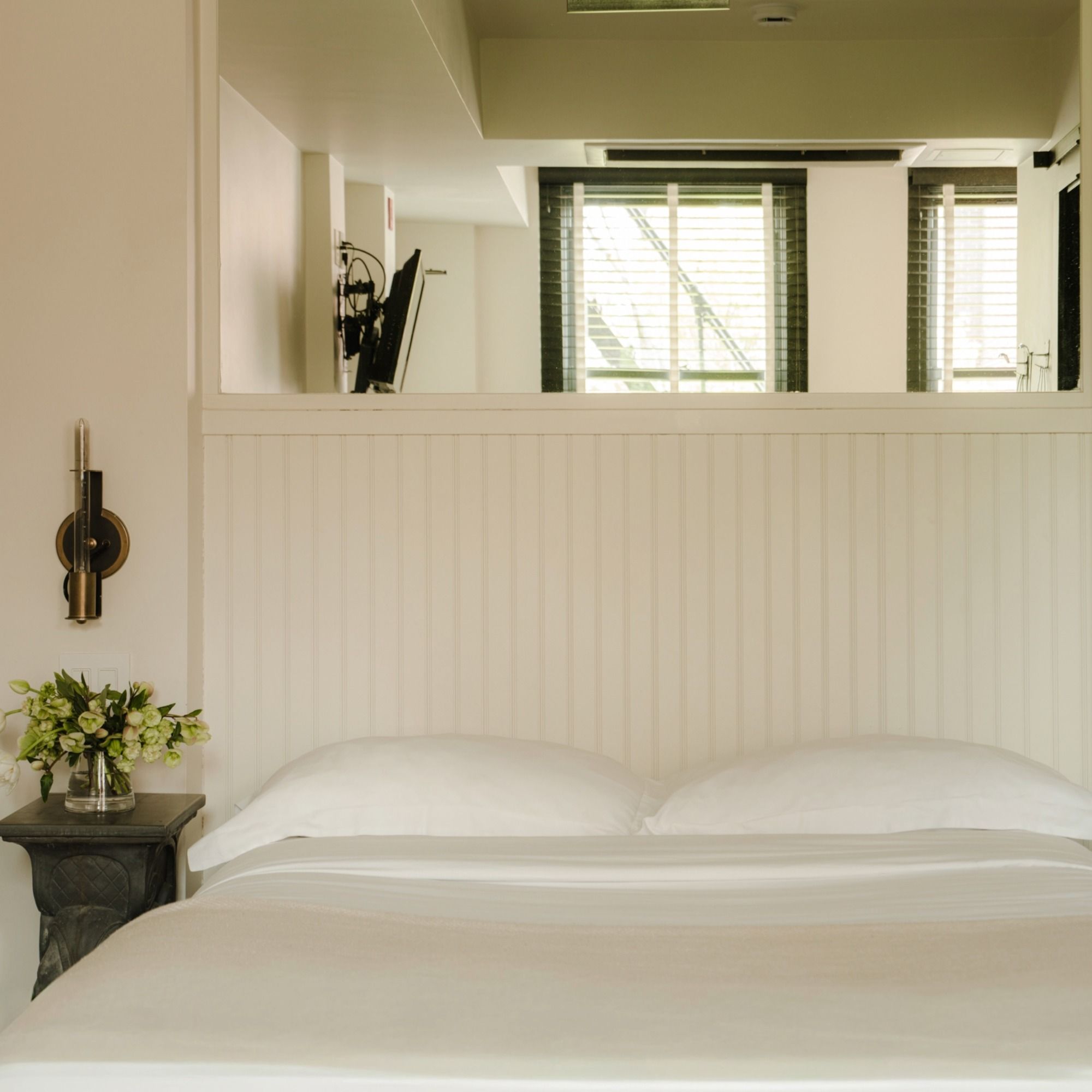 A cozy bedroom with white bedding, a nightstand with a vase of flowers, and a wall-mounted lamp. Sunlight filters through window blinds.