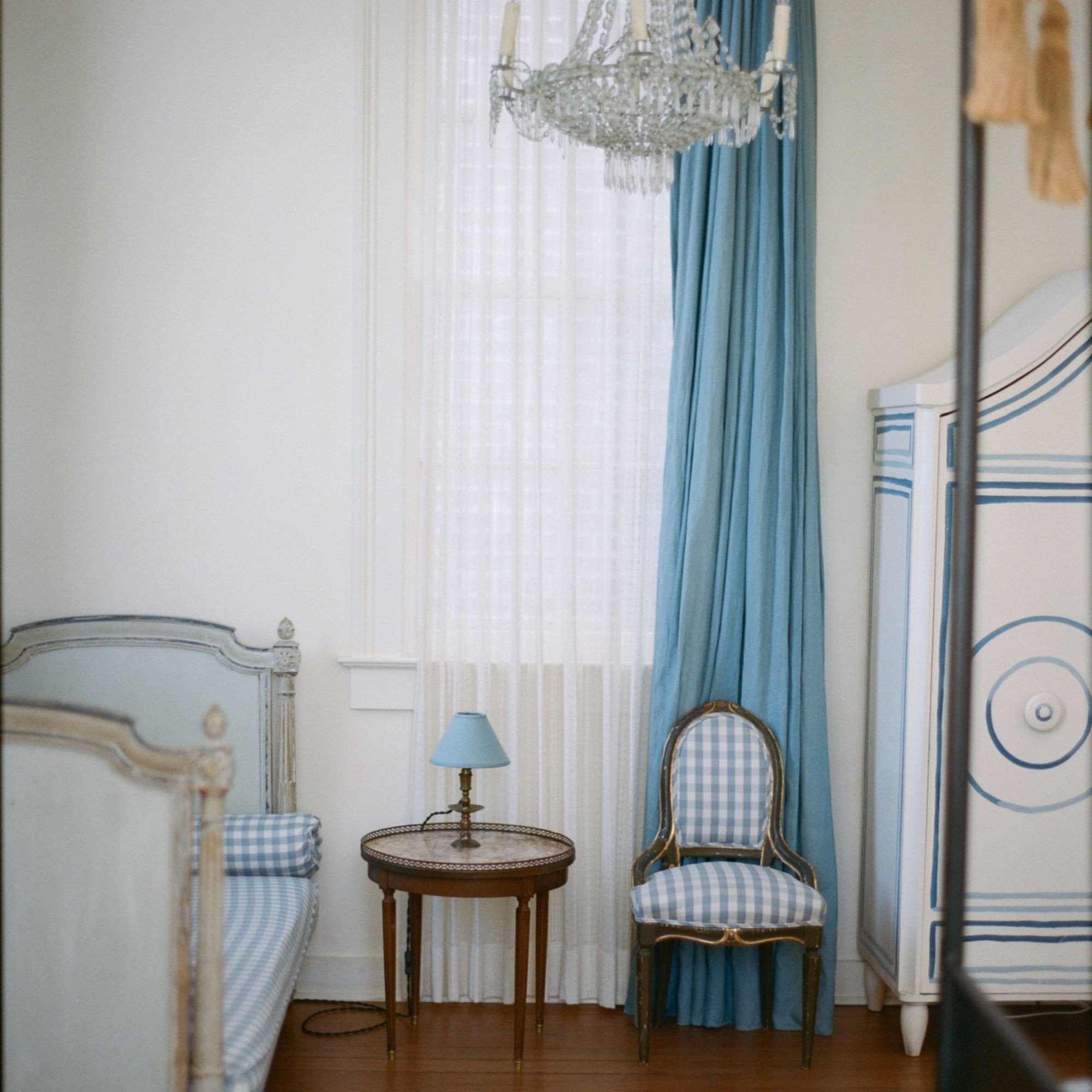 A vintage-style room with blue and white decor, featuring a bed, round table with a lamp, and a checkered chair by a window with sheer curtains.
