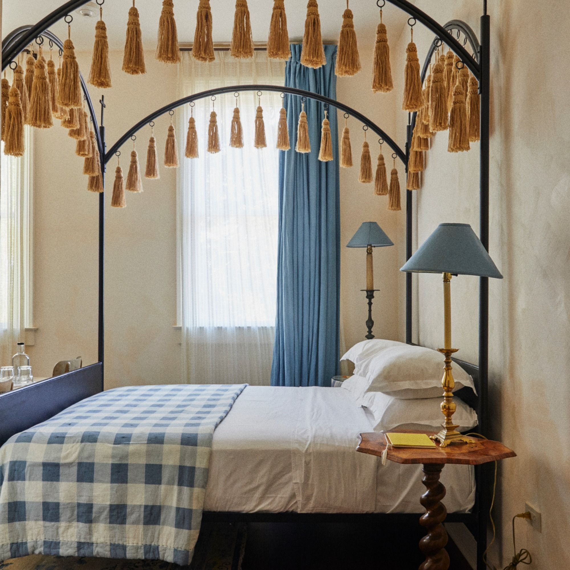 A cozy bedroom with a canopy bed featuring tassels, a blue-checkered blanket, blue curtains, and a wooden side table with matching lamps.