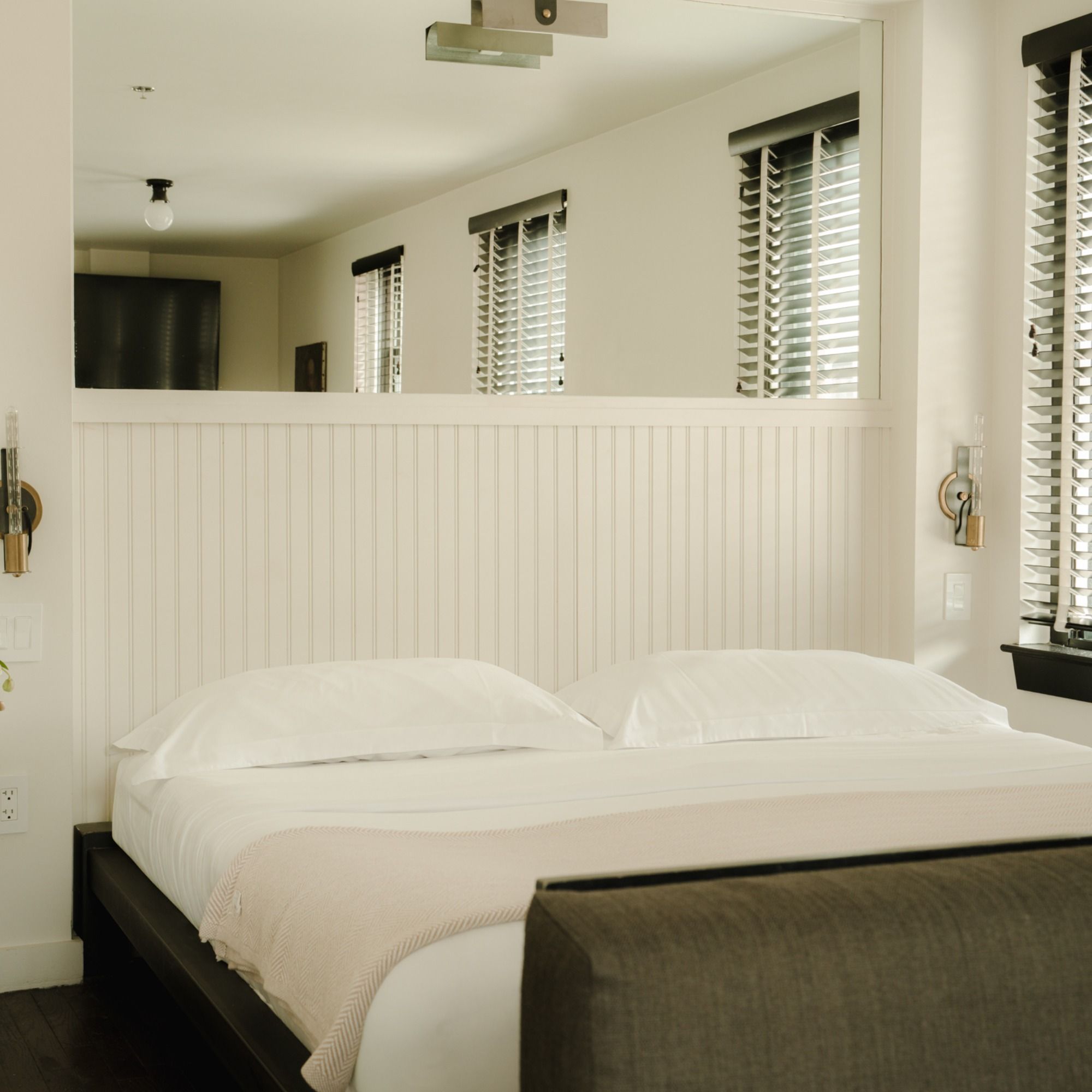 Modern bedroom with white bedding, a beige blanket, dark wooden bed frame, and large windows with black blinds, creating a serene ambiance.