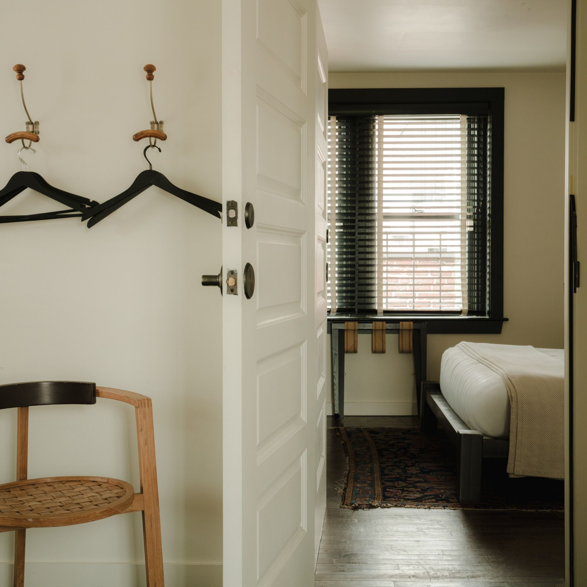 A minimalistic bedroom with a hanging chair, wood hangers on hooks, an open door, and a bed near a window with blinds.