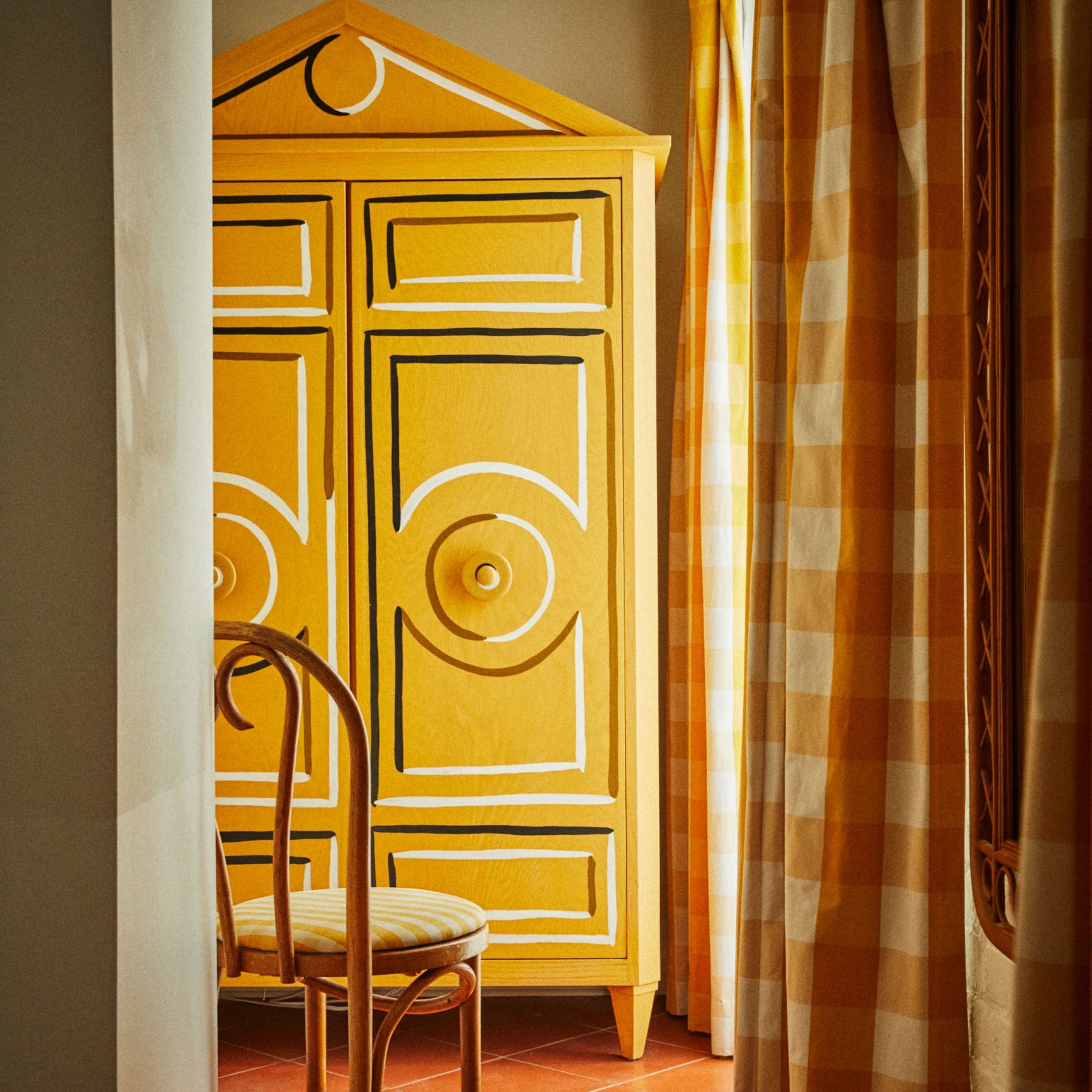Yellow wardrobe with decorative patterns beside yellow checkered curtains, and a wooden chair with a striped cushion in front.