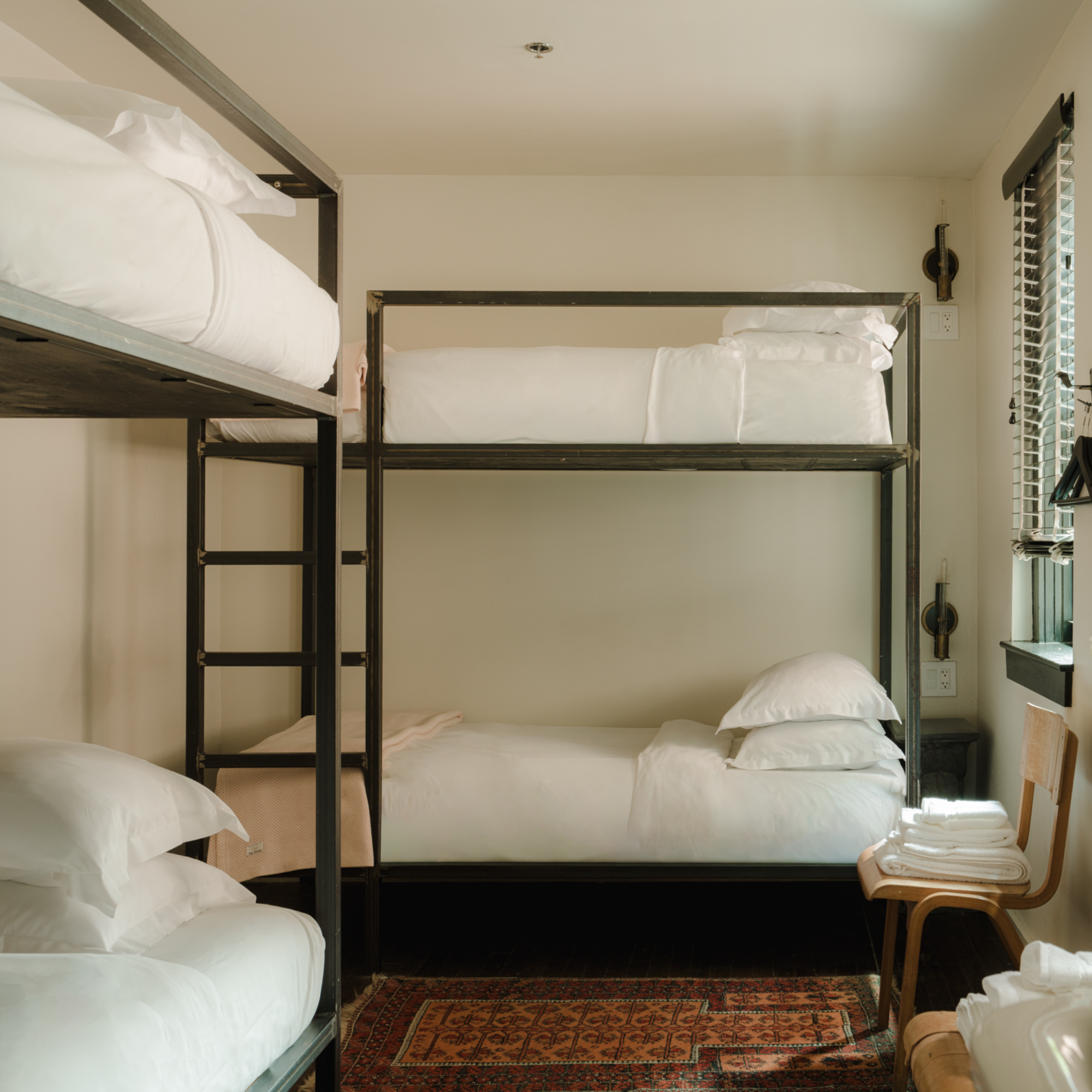 A cozy room with two bunk beds, white bedding, a wooden chair with folded towels, and a patterned rug on a dark floor. Natural light streams in.