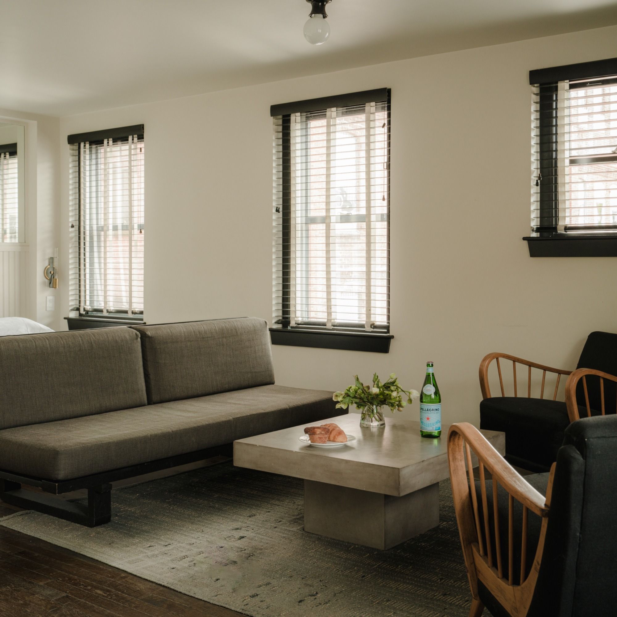 Modern living room with a gray sofa, concrete coffee table, wooden chairs, plant, pastry on a plate, mineral water bottle, and three windows with blinds.