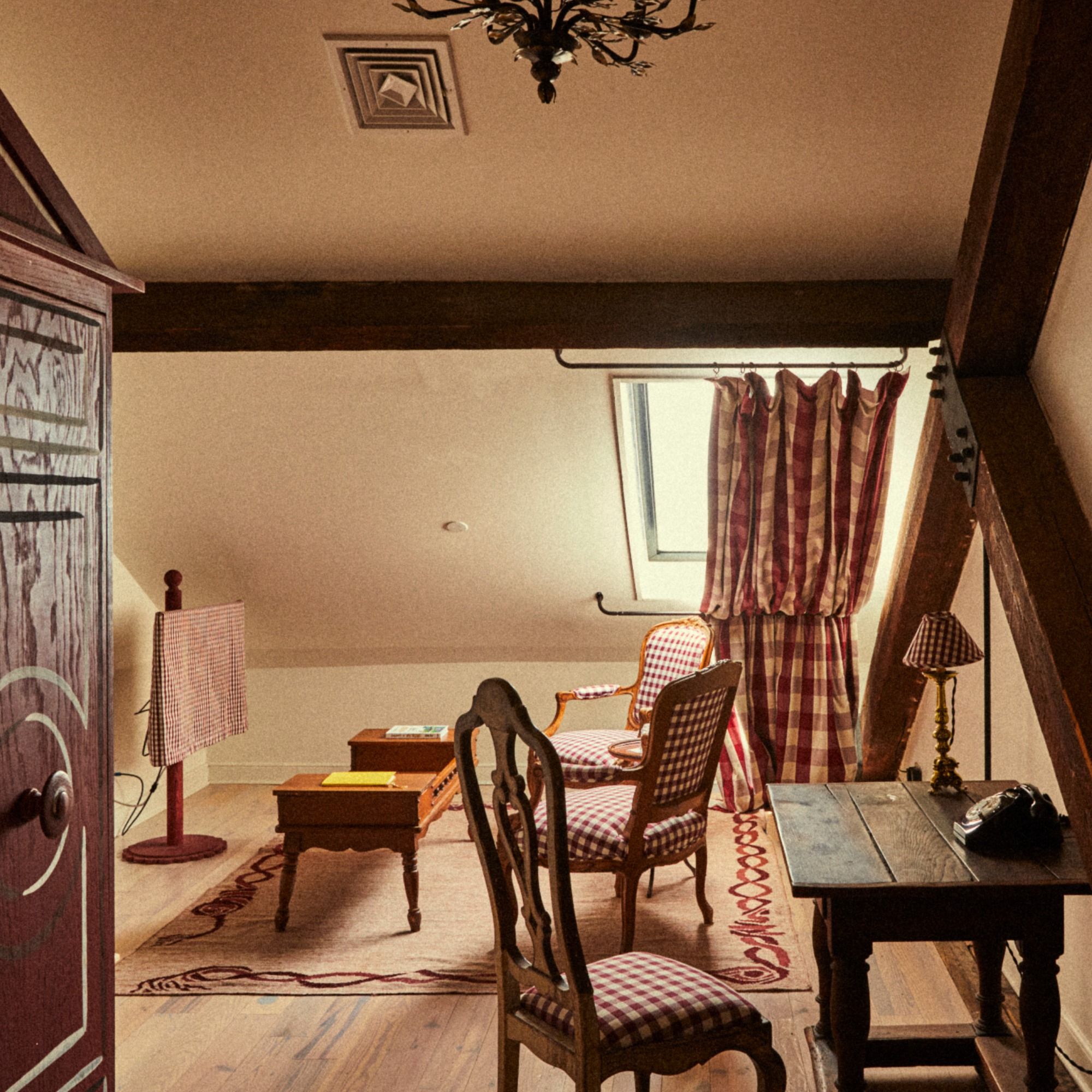Cozy attic room with wooden furniture, checkered upholstery, a skylight, and exposed beams. A small desk with a vintage phone is in the foreground.