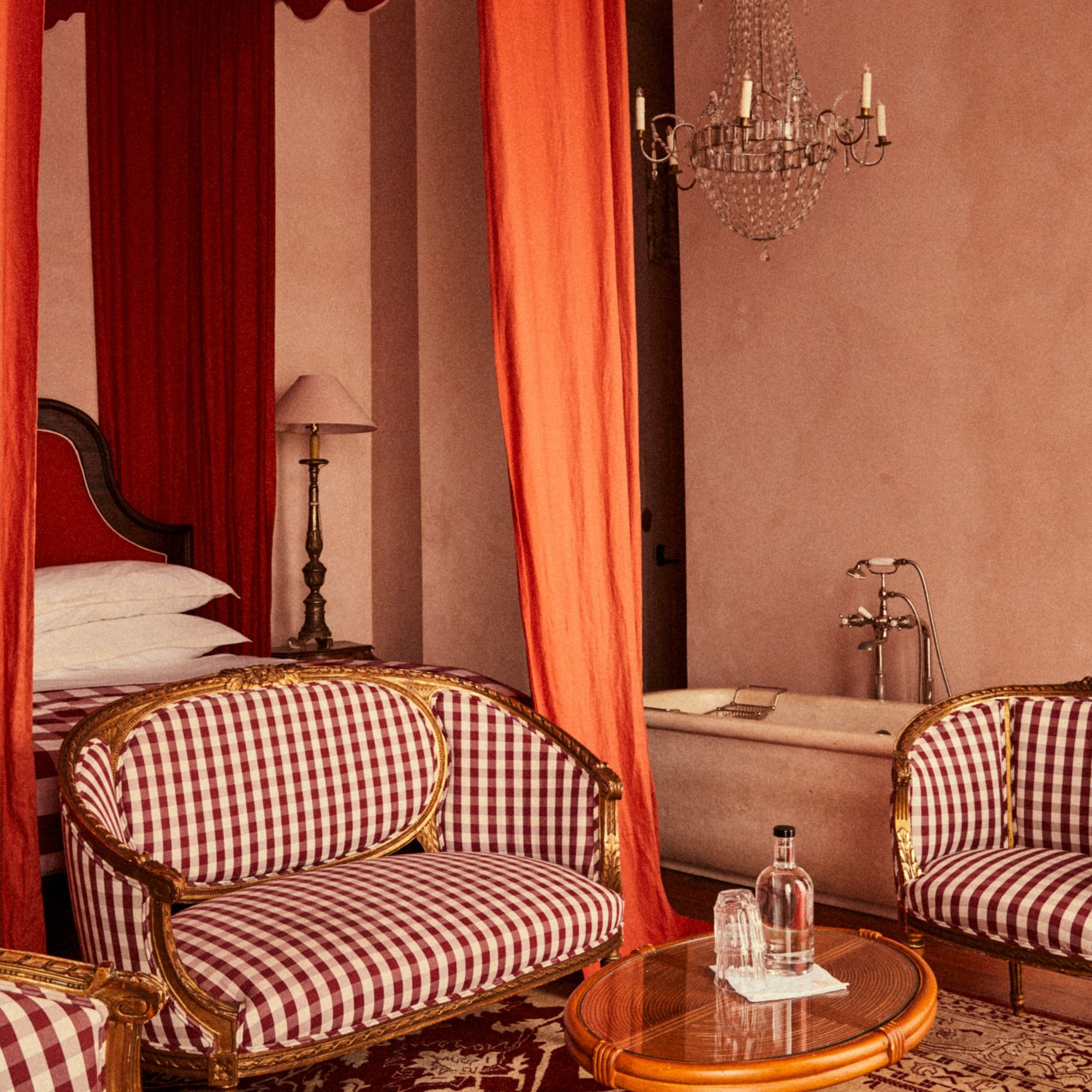 Luxurious room with red curtains, vintage striped chairs, ornate bed, chandelier, and bathtub. A small table holds a water bottle and glasses.