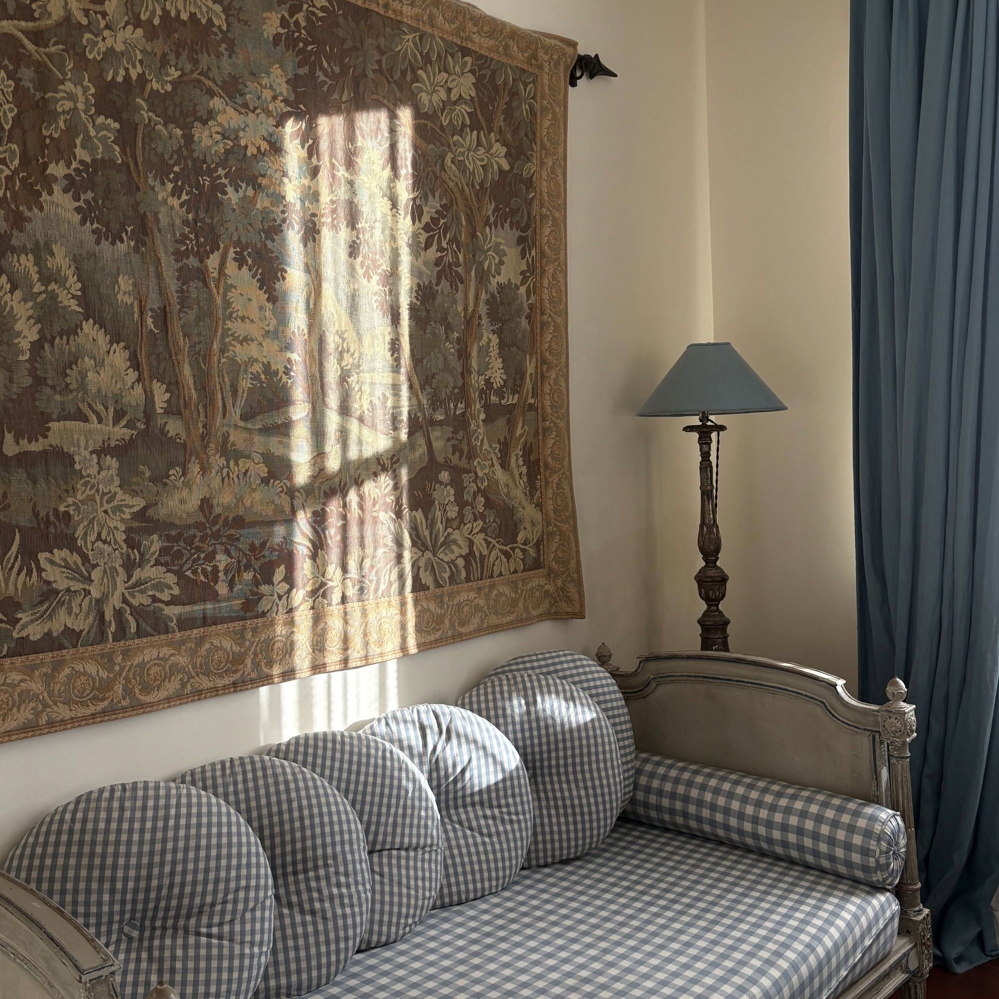 A cozy room features a checkered sofa with rounded cushions, a tapestry with a forest scene, and a decorative lamp beside a blue curtain.