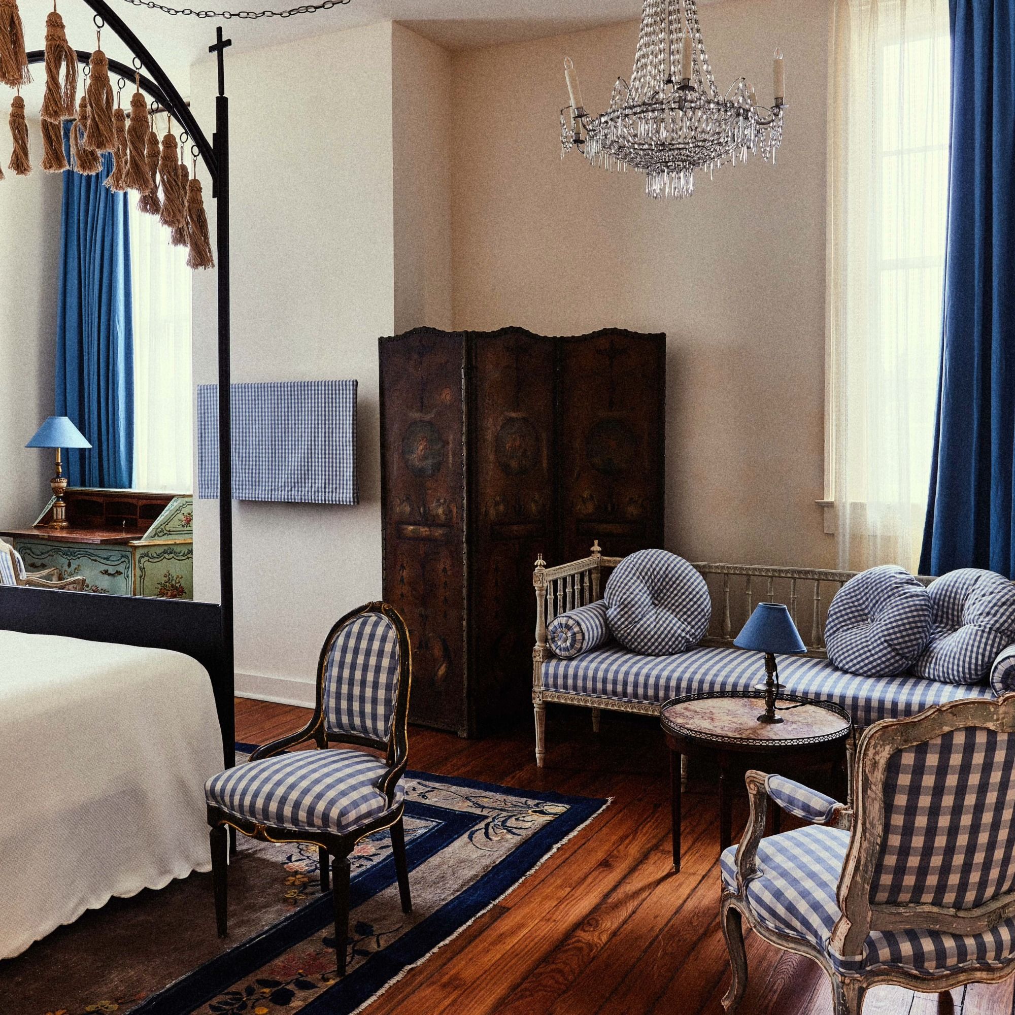 Elegant bedroom with blue gingham furniture, vintage armchairs, a decorative canopy bed, wooden floors, antique dresser, and a crystal chandelier.