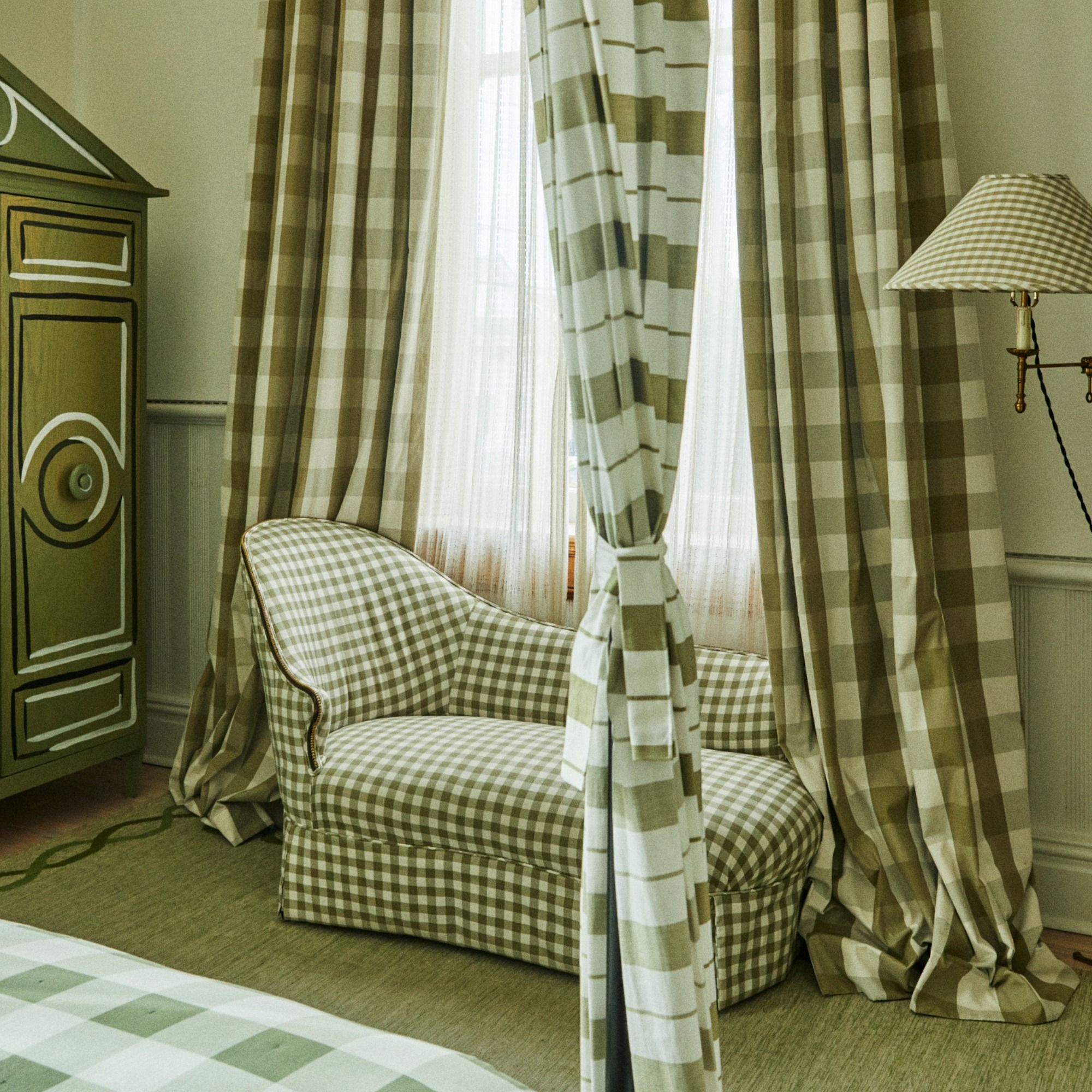 A cozy room with green checkered curtains, matching chaise lounge, green armoire, and a checkered floor lamp by a window with sheer drapes.