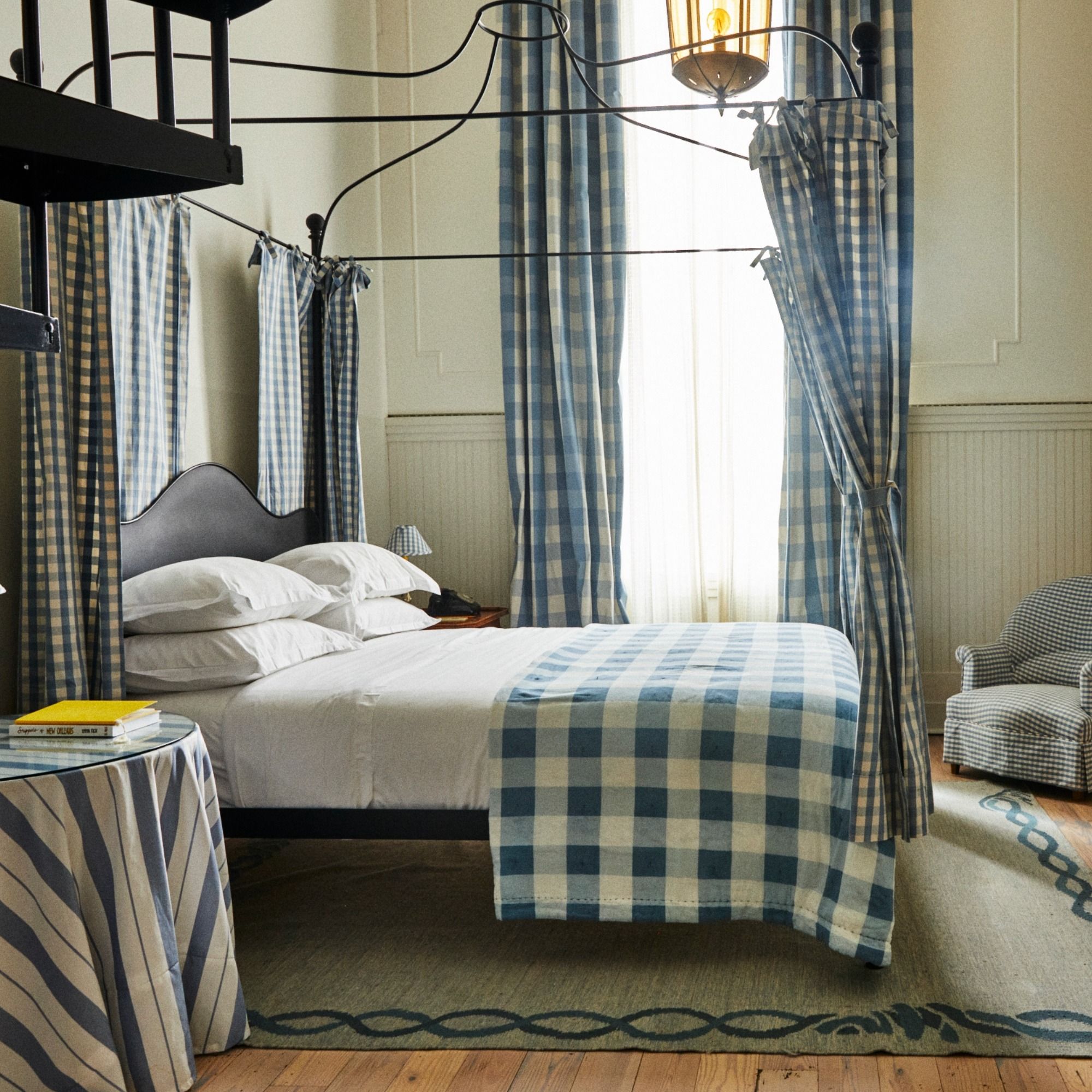 Bedroom with a four-poster bed dressed in blue and white checkered linens. Matching curtains, an armchair, and a lamp add to the cozy decor.