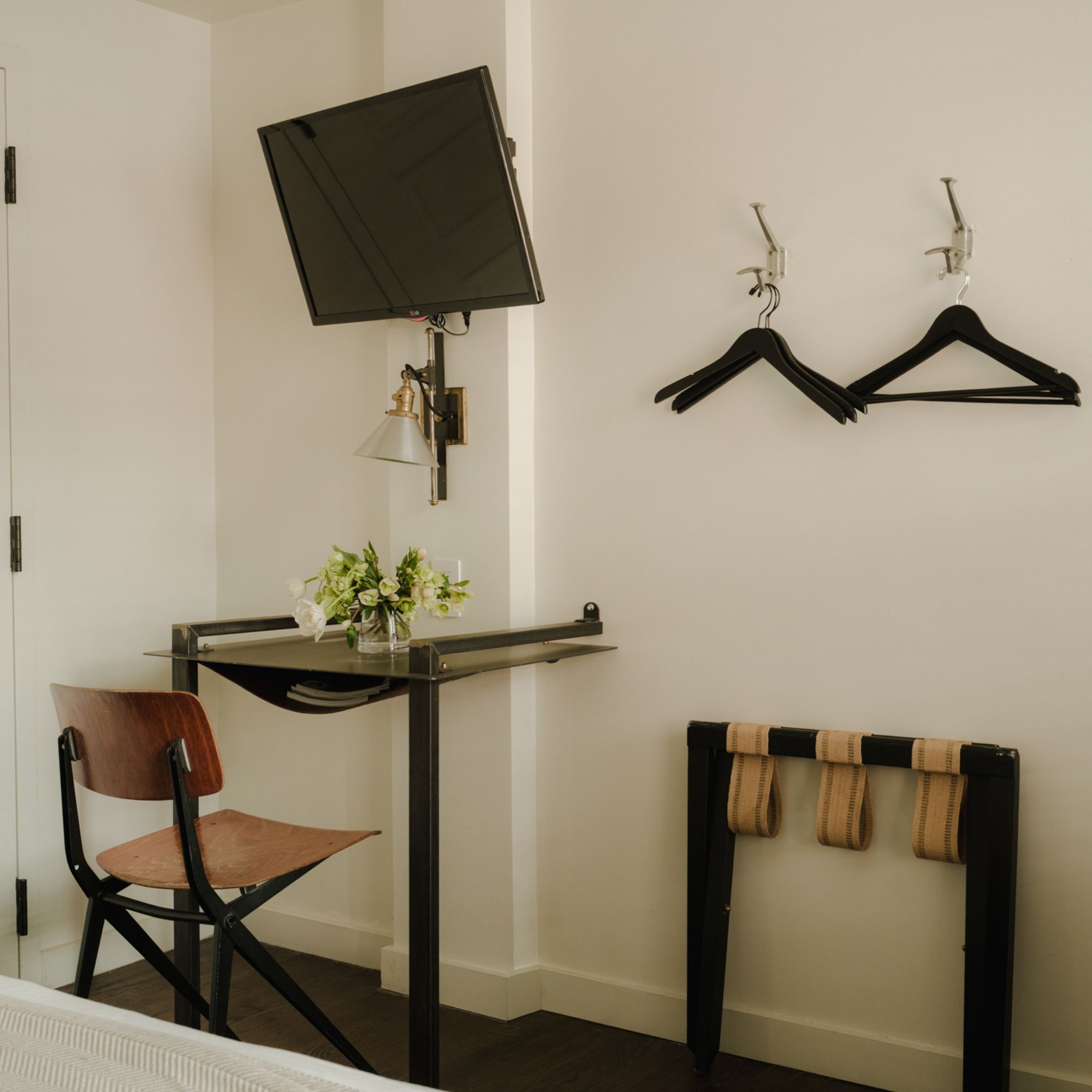 Small hotel room corner with a wall-mounted TV, metal desk holding flowers, wooden chair, coat hangers, and luggage rack on dark flooring.