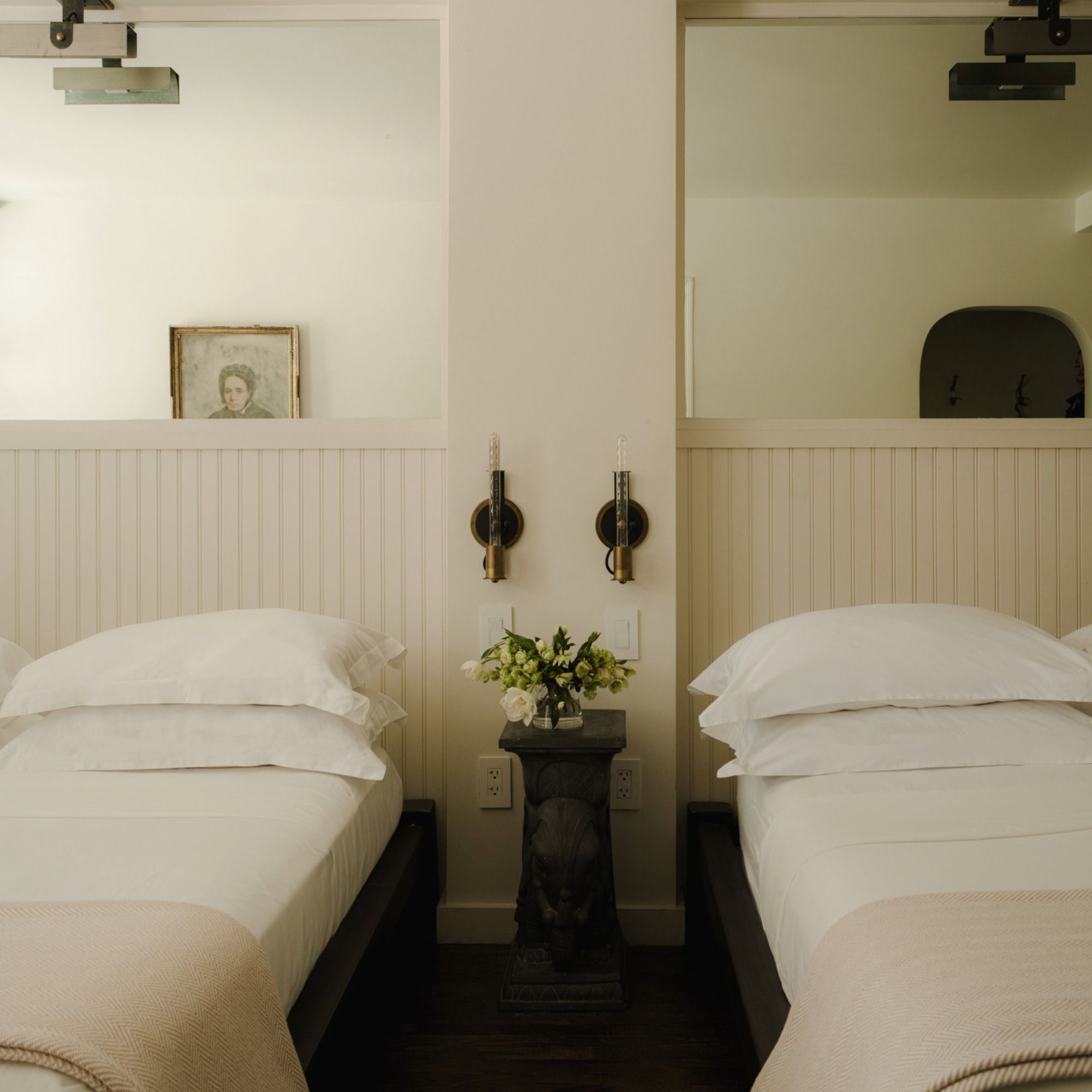 Two neatly made twin beds with white linens, separated by a table with flowers. Mirrors and sconces above, and a portrait in the background.