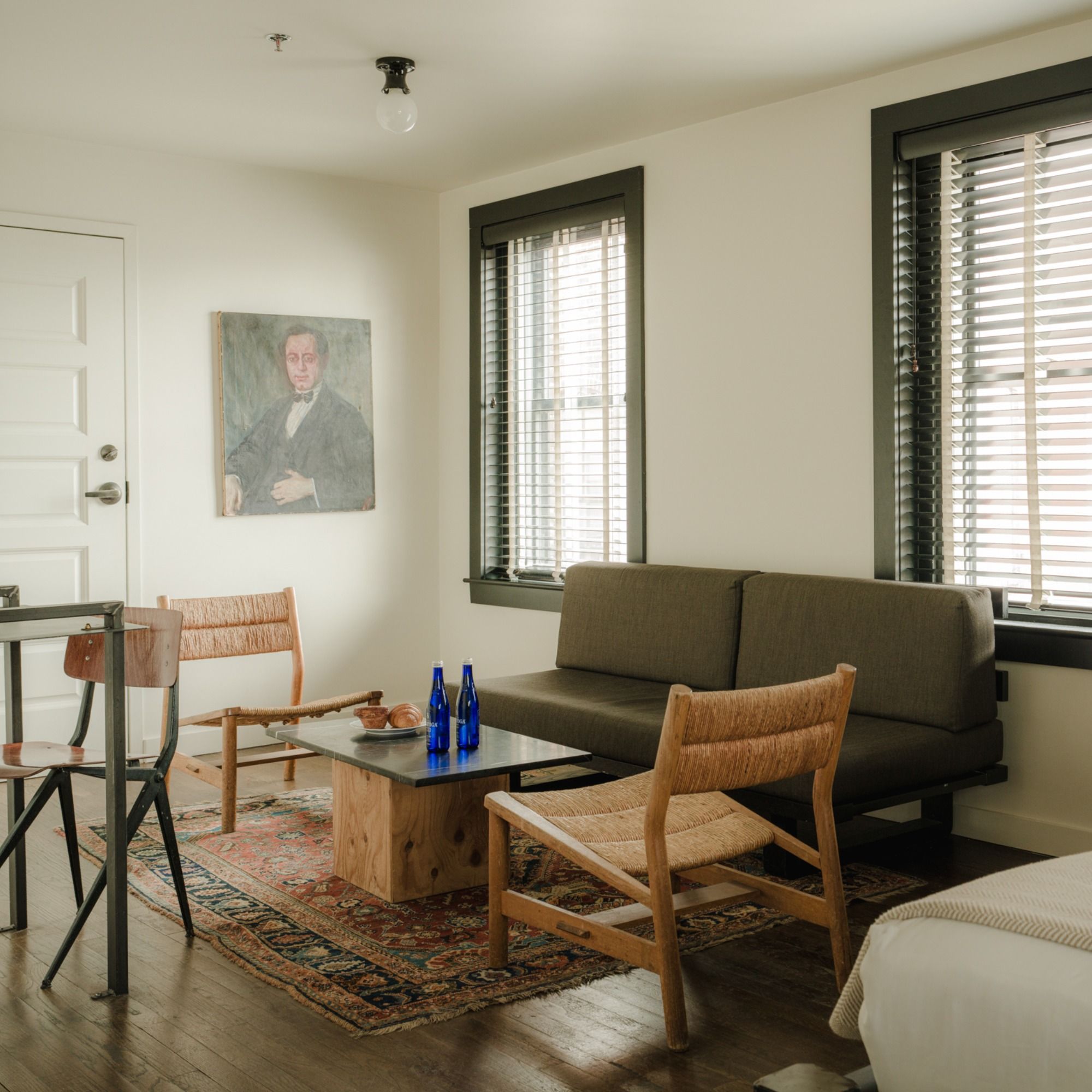 Cozy living room with a gray sofa, wooden chairs, a coffee table, and a rug. A painting hangs on the wall, and sunlight filters through the blinds.