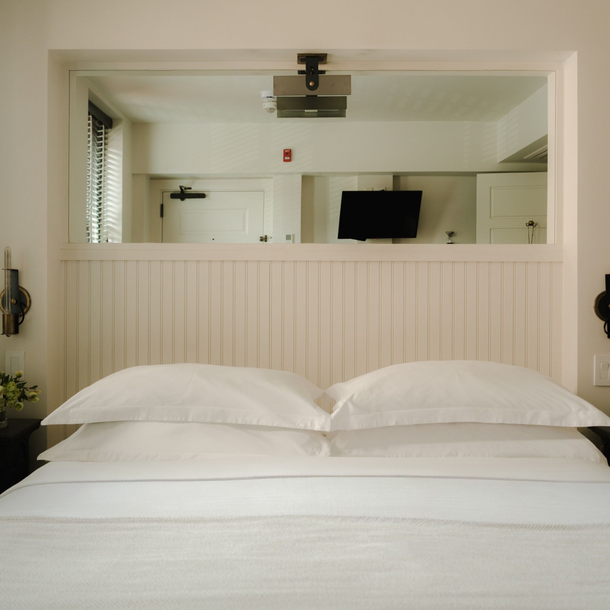 A cozy bedroom with neatly arranged white pillows and bedding, panelled wall, and a window showing an adjoining room with a TV and door.