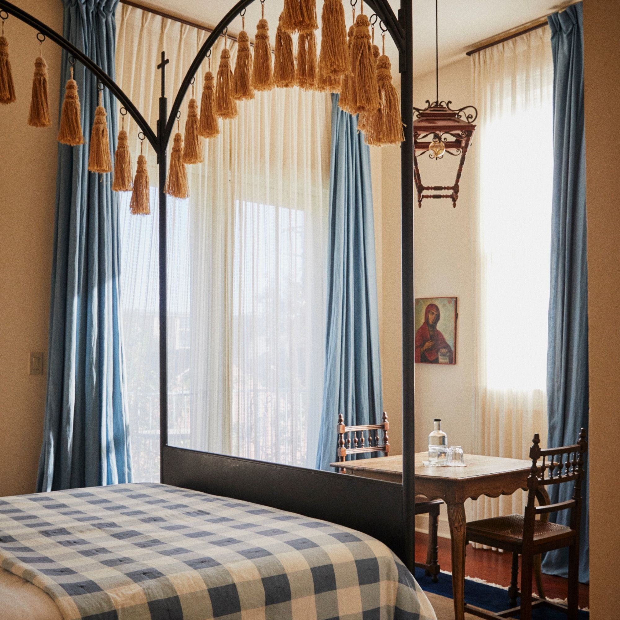 A cozy bedroom with a canopy bed and blue plaid blanket, tassels, a table with a water carafe and glasses, and light streaming through blue curtains.