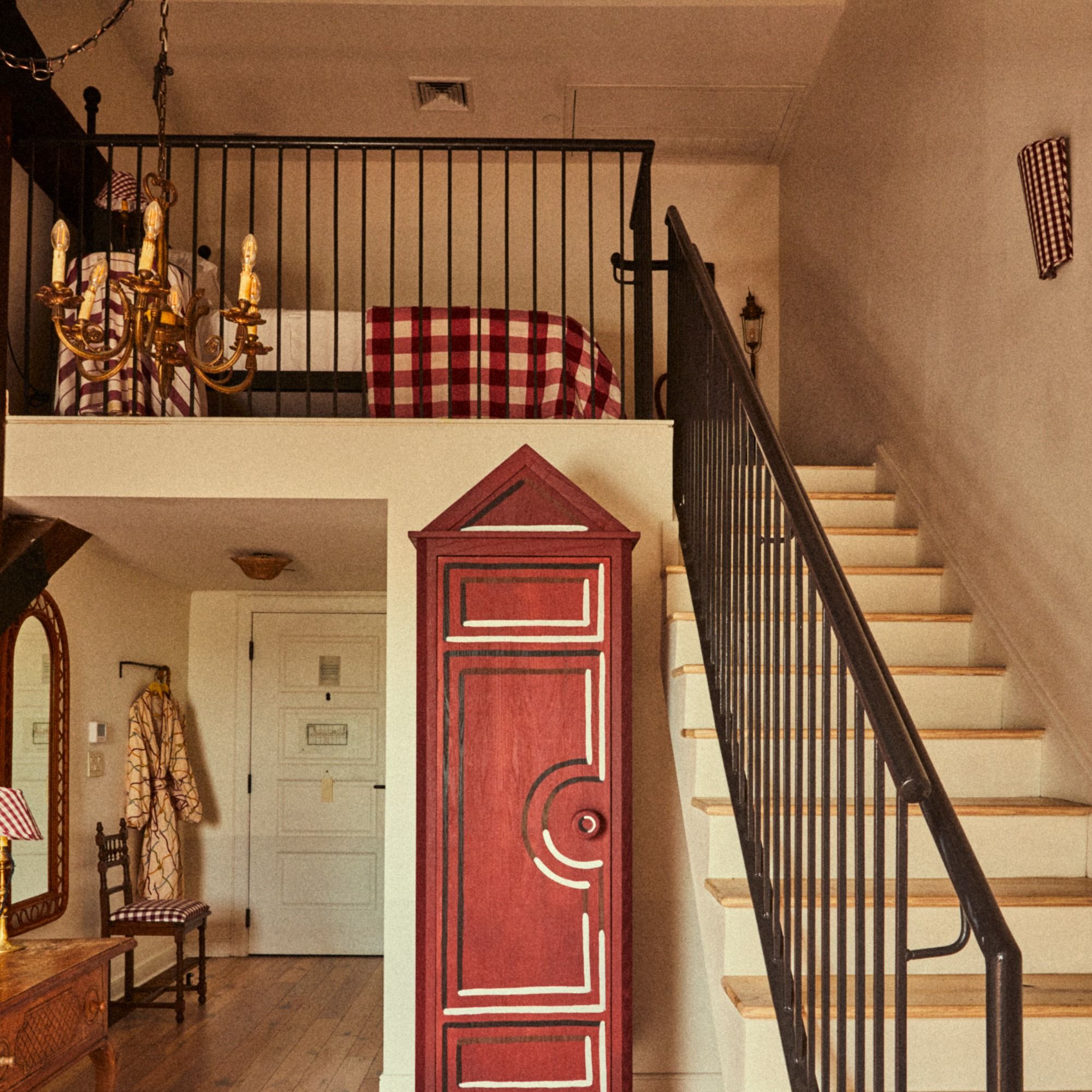 Cozy loft with a red wardrobe, checkered bedspread above, wooden chair, decorative chandelier, and a staircase leading to the upper level.
