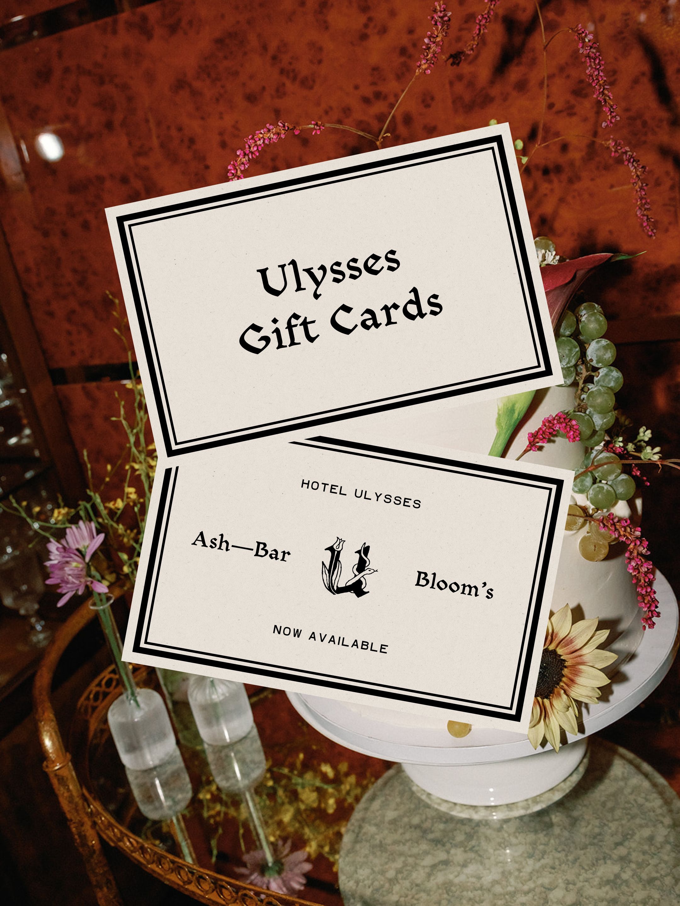 Two Ulysses gift cards atop a cake stand surrounded by flowers and grapes on a mirrored table.
