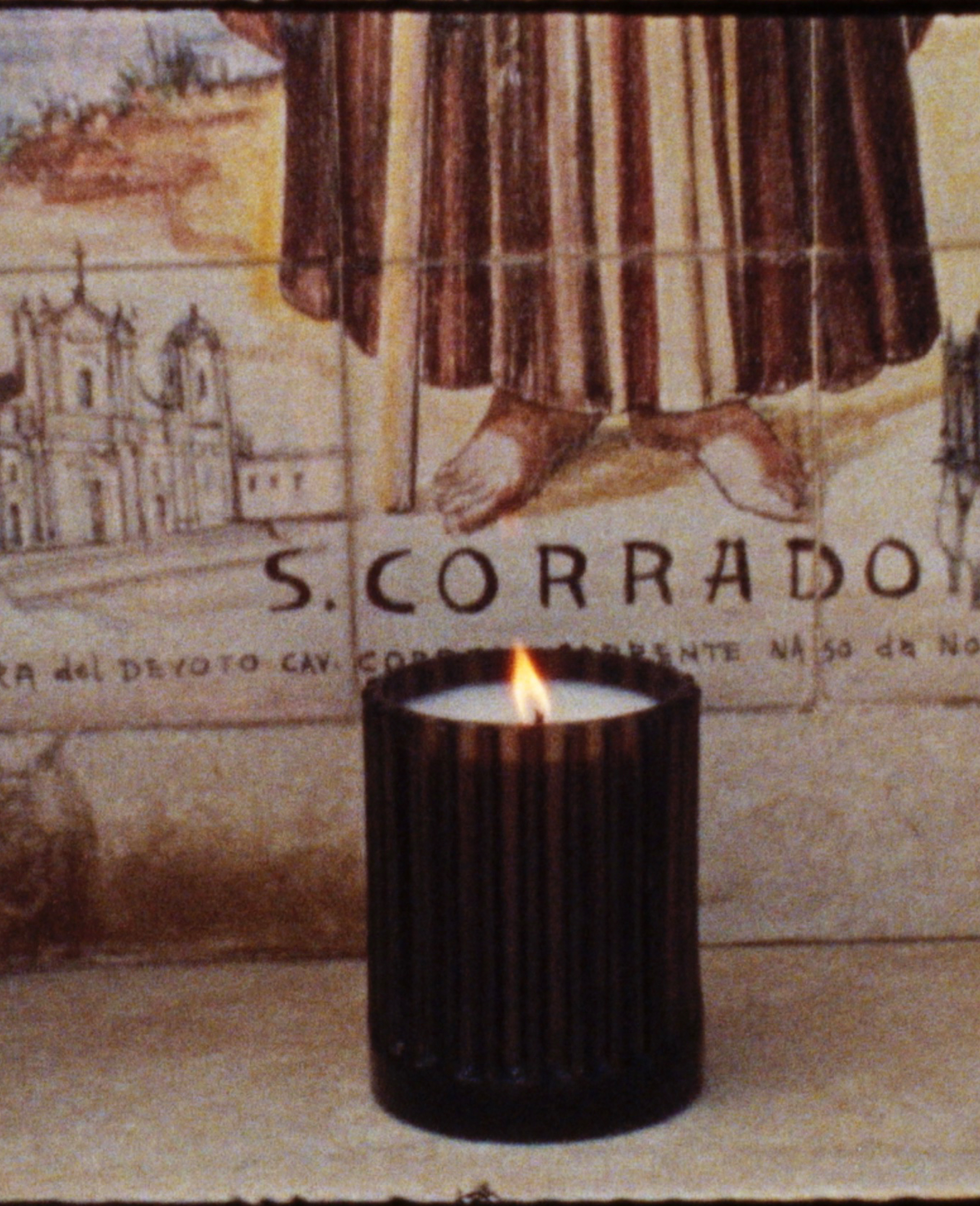 A lit candle in a dark holder sits in front of a tile mural depicting a religious figure and the text "S. CORRADO."
