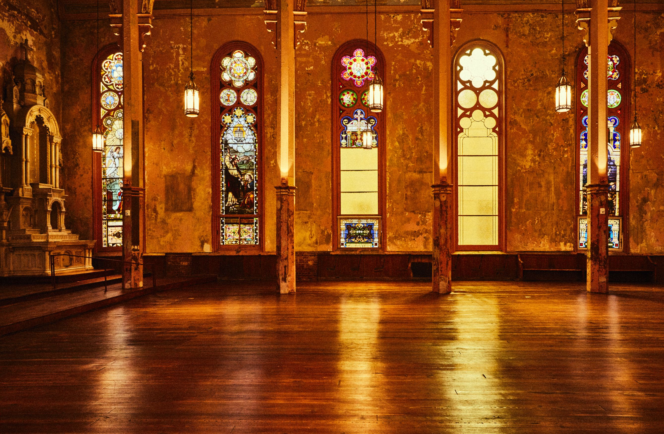 Warm-lit interior of a historic church with stained glass windows, wood flooring, and hanging lamps, creating a serene atmosphere.
