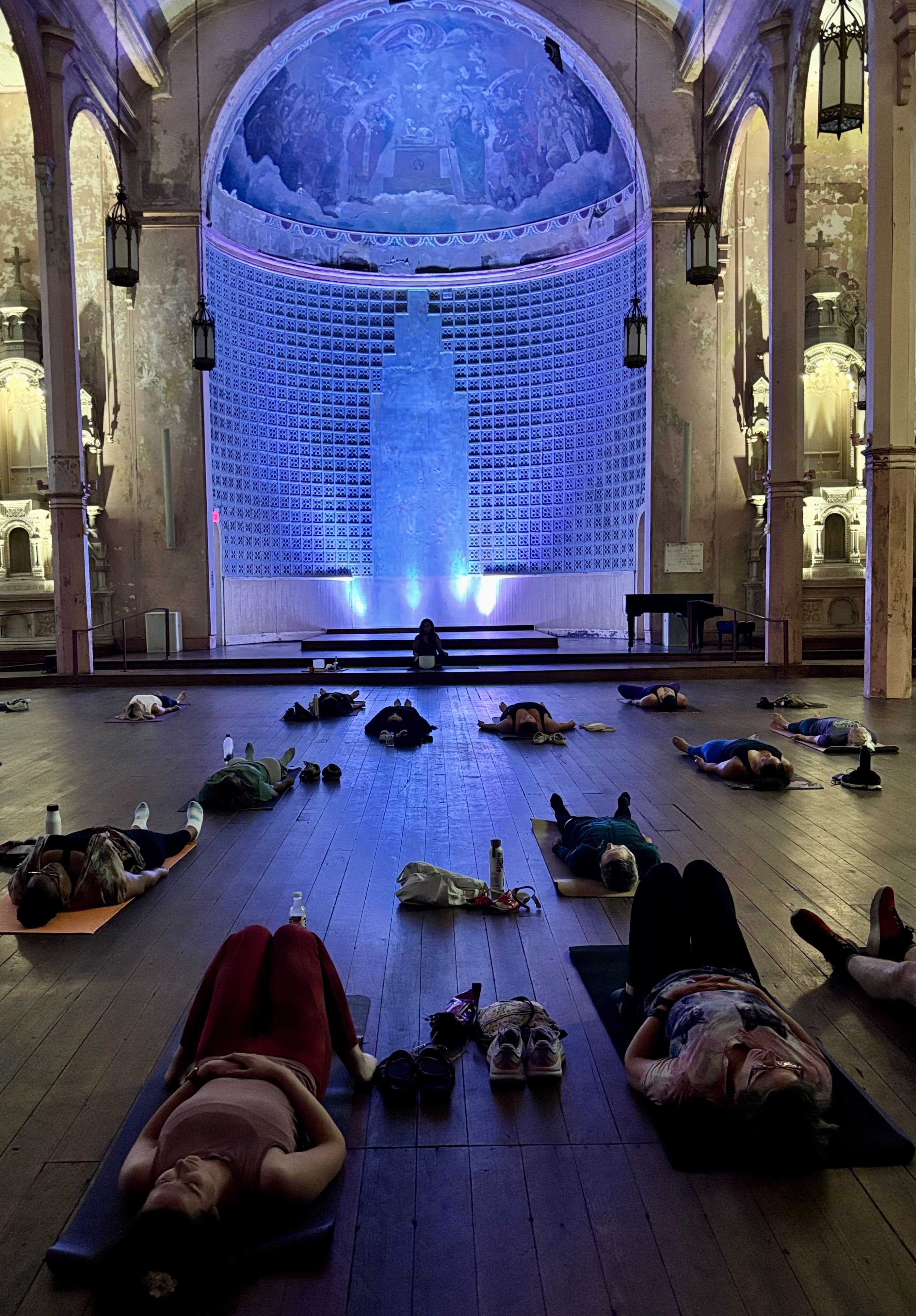 People lying on yoga mats in a dimly lit, ornate hall with a painted dome and soft colored lights on the stage.