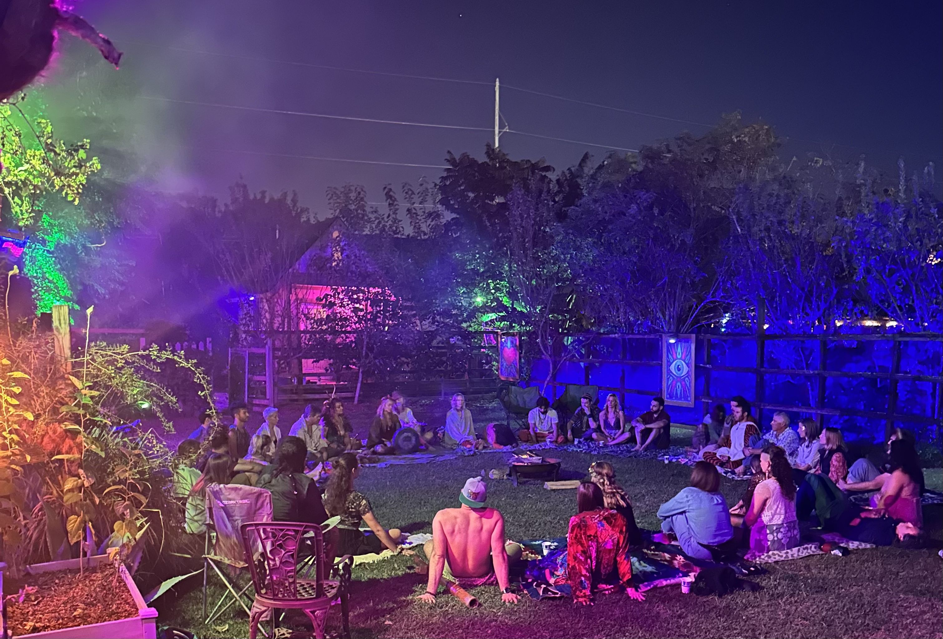 A group of people sit around a bonfire in a garden at night, surrounded by colorful lights and trees, with a clear starry sky above.