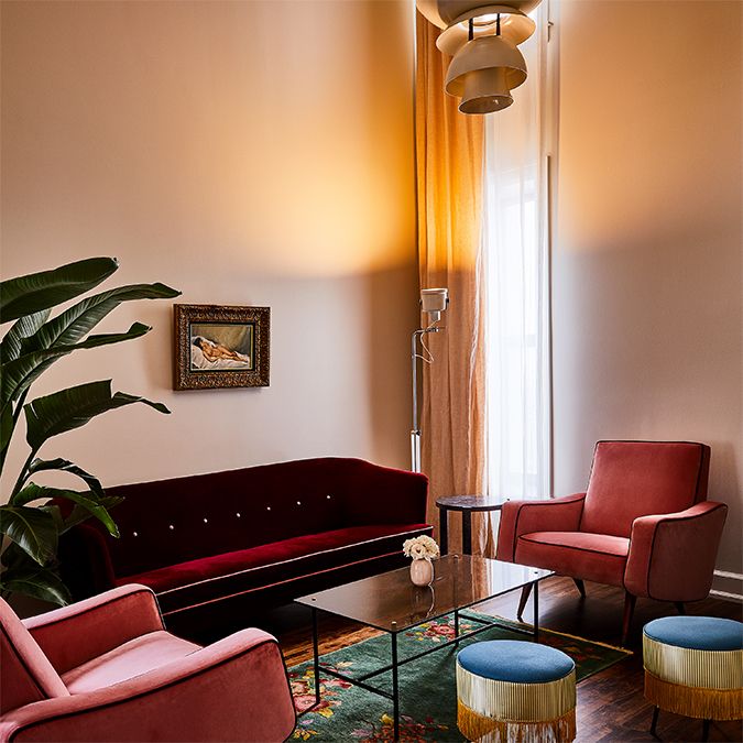 Stylish living room with pink chairs, a red sofa, large plant, framed painting, and soft lighting. A tall window with sheer curtains brightens the space.