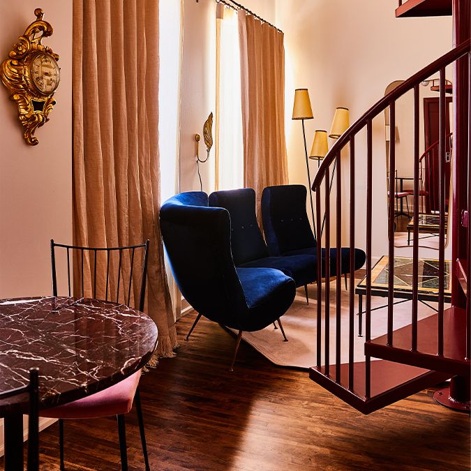 Cozy room with a spiral staircase, velvet blue chairs, a round marble table, and tall curtains. Elegant decor and warm lighting.