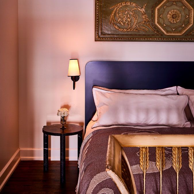 Cozy bedroom with a navy headboard, decorative pillows, a wall-mounted lamp, and a nightstand with a small lamp and flower vase.