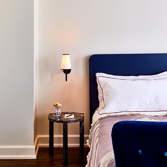 A cozy bedroom corner featuring a blue upholstered bed, a small round side table with a vase of flowers, and a wall-mounted sconce.