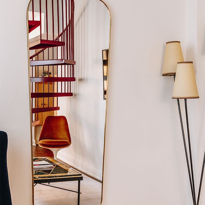 Tall mirror reflecting a room with a red spiral staircase, an orange chair, a green table, and a floor lamp with three beige lampshades.