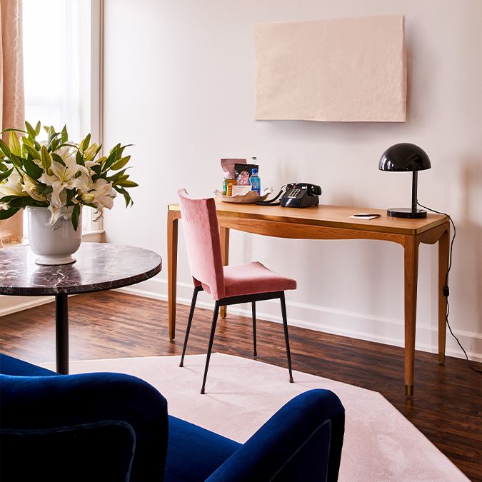 A stylish workspace with a wooden desk, pink chair, black lamp, telephone, and flowers on a side table. Cozy and elegant setting.