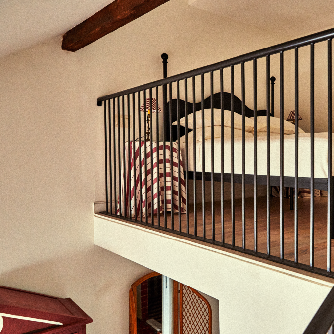 Loft bedroom with black metal railing, wooden beam ceiling, and a bed featuring a red and white checkered blanket, next to a striped lamp.