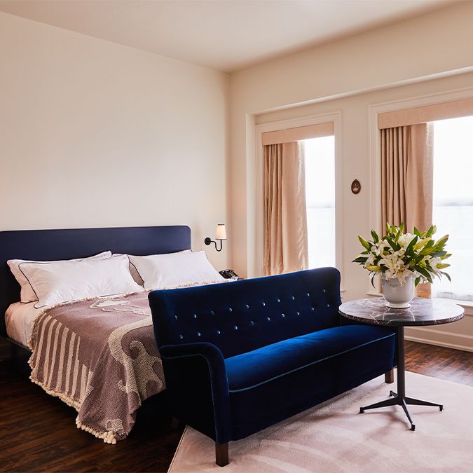 A cozy bedroom with a navy bed, matching velvet loveseat, round table with lilies, and beige curtains over large windows.