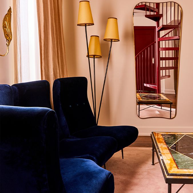 Elegant living room with deep blue armchairs, tall lamps, a decorative mirror, and a colorful table. A spiral staircase is reflected in the mirror.